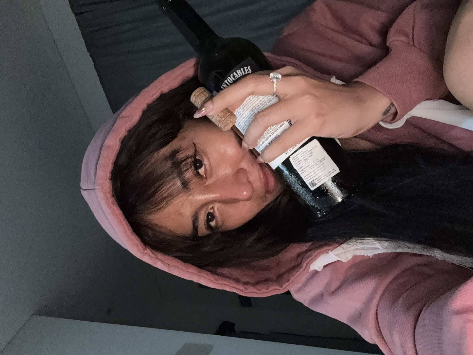 A close-up, dimly lit selfie captures a person, likely female, in a relaxed pose, holding a wine bottle near their face. The individual has long, dark hair, with bangs falling across their forehead, and is wearing a pink hooded sweatshirt. Their right hand, adorned with a prominent ring featuring a large, clear stone, holds a dark glass wine bottle. The cork from the bottle is also visible, held close to their cheek.
The person looks directly into the camera with a soft, somewhat ambiguous expression. The lighting is dim, suggesting an indoor evening or nighttime setting, with reflections from an unseen light source visible on the bottle and the person's skin.
Visible text on the wine bottle label includes the brand name "INTOCABLES," along with phrases like "PRODUCED & BOTTLED B," "ARGENTINA WINE ARGENTINA'S NATIONAL BEVERAGE," "CONTAINS SULPHITES," and "ALC. 13.5%/VOL." Other smaller, partially legible text on the label describes tasting notes such as "aromas" and "caramel." The scene conveys a casual and intimate moment.