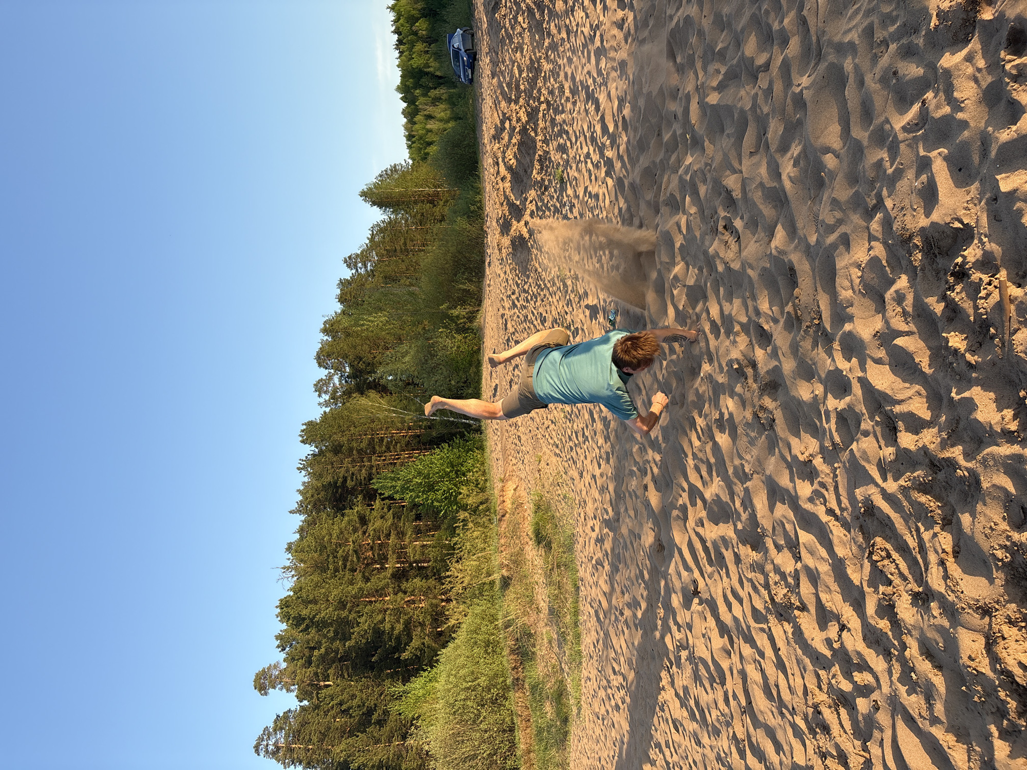 A man is lying prone on a sandy beach, tossing sand into the air. He is wearing a short-sleeved blue-green shirt and shorts. The beach is adjacent to a forest of evergreen trees. A vehicle is visible in the distance, parked near the edge of the forest. The sky is clear blue. The location is Chaikovsky, Russia.