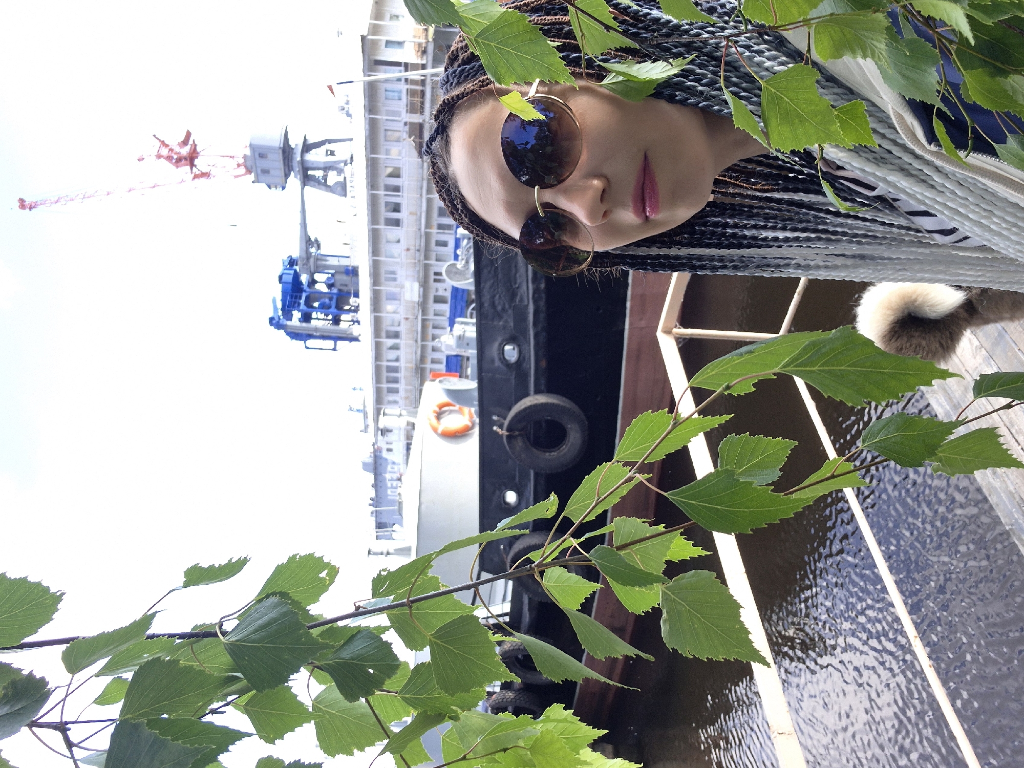 In Chaikovsky, Russia, a woman with dark braided hair and round sunglasses stands on a pier, partially obscured by green leaves in the foreground. She is looking to her right. Behind her, a large dark-hulled ship with life preservers and tires along its side is docked in a body of water. In the background, industrial cranes and a multi-story building with many windows are visible, suggesting a port or industrial area. The sky is bright and appears overcast or hazy.