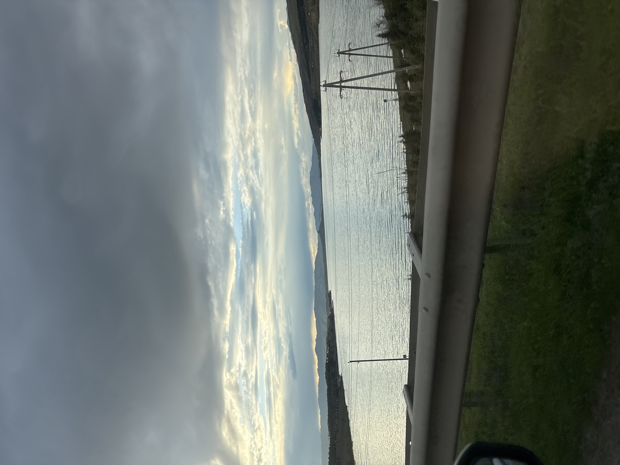 The image shows a view from a vehicle, possibly a car, looking out over a body of water, likely a lake or river. A guardrail is visible on the right side of the frame, with green vegetation extending beyond it. Power lines and pylons are visible along the opposite shore of the water, indicating some level of infrastructure. The sky is partially obscured by clouds.
Without more information, it's impossible to conclusively determine the exact location of this scene in Tbilisi, Georgia.