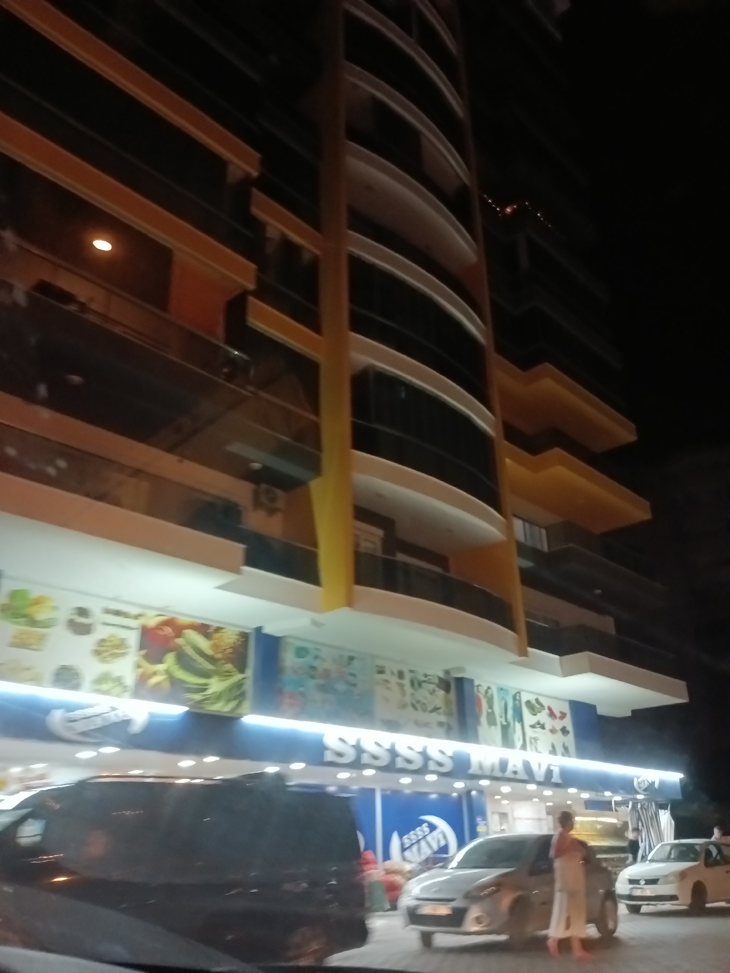 In Alanya, Turkey, at night, a multi-story building with modern architecture is illuminated. The building features curved balconies with dark glass and prominent yellow vertical accents. Below the building, a storefront with a bright blue sign reading "SSSS MAVI" is visible. This sign is illuminated by white lights. Various images of clothing and footwear are displayed on the wall above the sign. In front of the store, two cars are parked. A silver hatchback is in the foreground, and a white sedan is to its right. A person, appearing to be a woman wearing a white towel or robe and pink slippers, stands next to the silver car. The ground in front of the store appears to be a paved area, possibly a sidewalk or parking lot. The overall scene suggests a commercial area in Alanya at night.