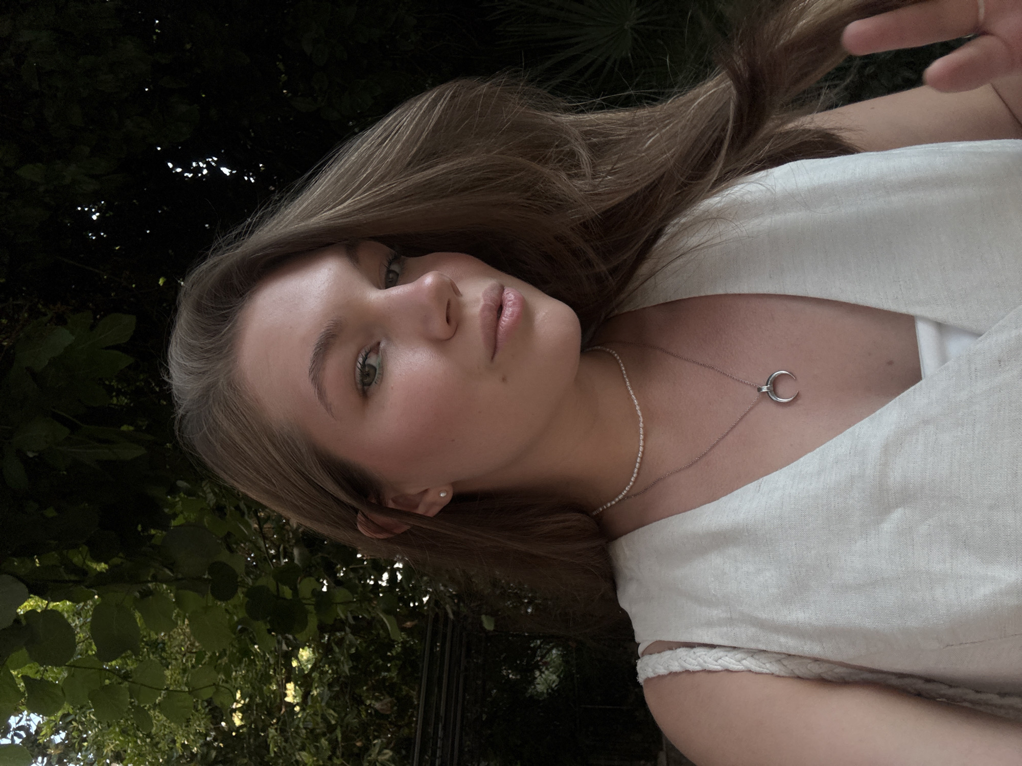 A young woman with long, light brown hair and light green eyes is shown in a close-up, outdoor portrait. She is wearing a light-colored, sleeveless top over a white camisole, and a delicate pearl necklace with a crescent moon pendant. The background is filled with lush green foliage, suggesting a garden or park setting. The lighting is soft, with some areas of shadow, indicating it might be late afternoon or early evening. The overall atmosphere is casual and relaxed. While the image itself does not provide specific geographical markers, it evokes a sense of natural beauty and tranquility that could be found in numerous outdoor locations, including the scenic areas of Budva, Montenegro.