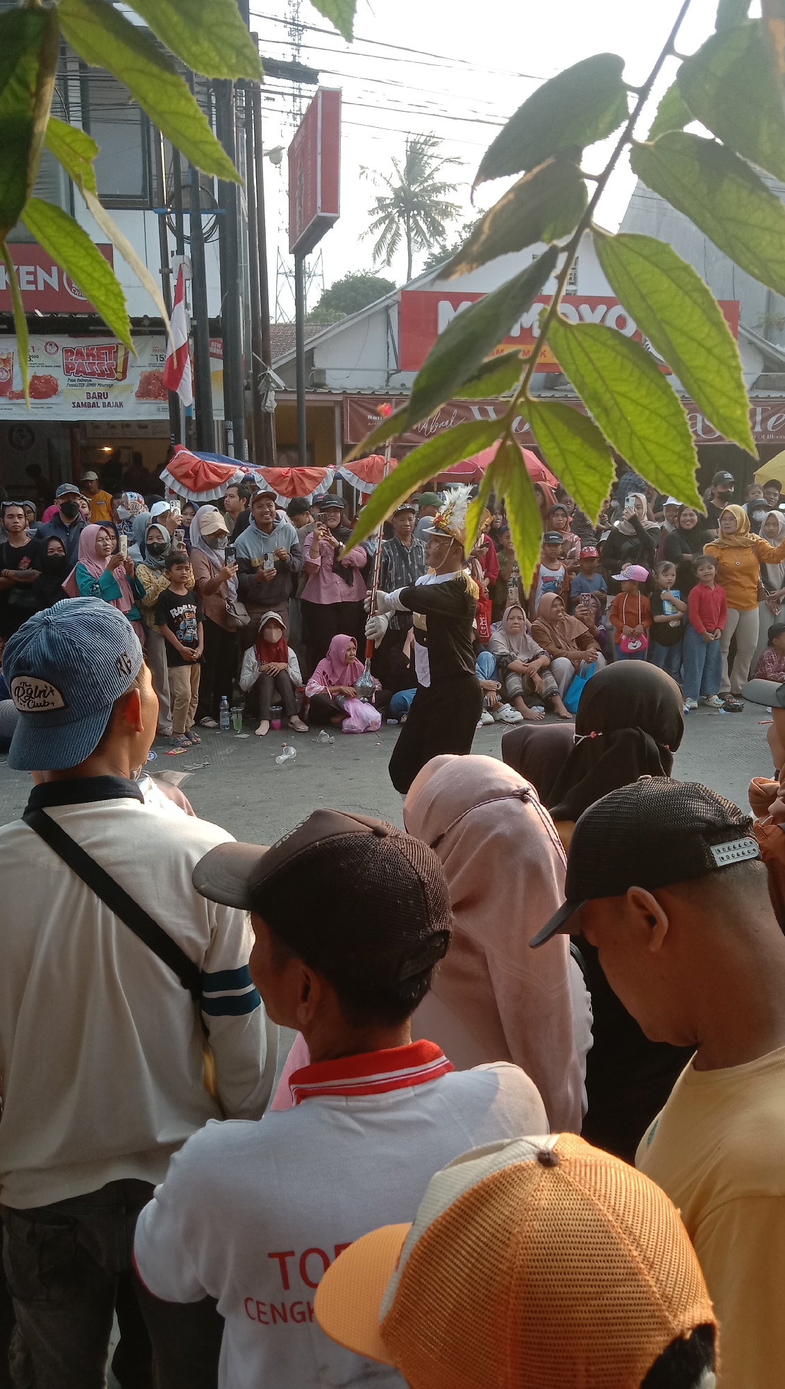 A street performance is taking place in front of a row of shops in Jatisawit, Indonesia. The event appears to be a parade or festival, with a crowd gathered to watch. A young person in a black and white uniform, wearing a hat with a plume, is at the center of the activity, holding a baton. They are performing a baton-twirling routine. The crowd consists of people of various ages, many of whom are watching the performance. Some are holding up their phones to take pictures or record videos. In the background, there are shop fronts with signage, including one that reads "MOMOYO". Red and white streamers are strung across the street, suggesting a celebratory atmosphere. The lighting suggests it is daytime, possibly late afternoon. The presence of palm trees in the background indicates a tropical climate. Some of the people in the foreground are wearing hats, and some of the women are wearing headscarves. The overall scene is one of a community gathering for a local event.
