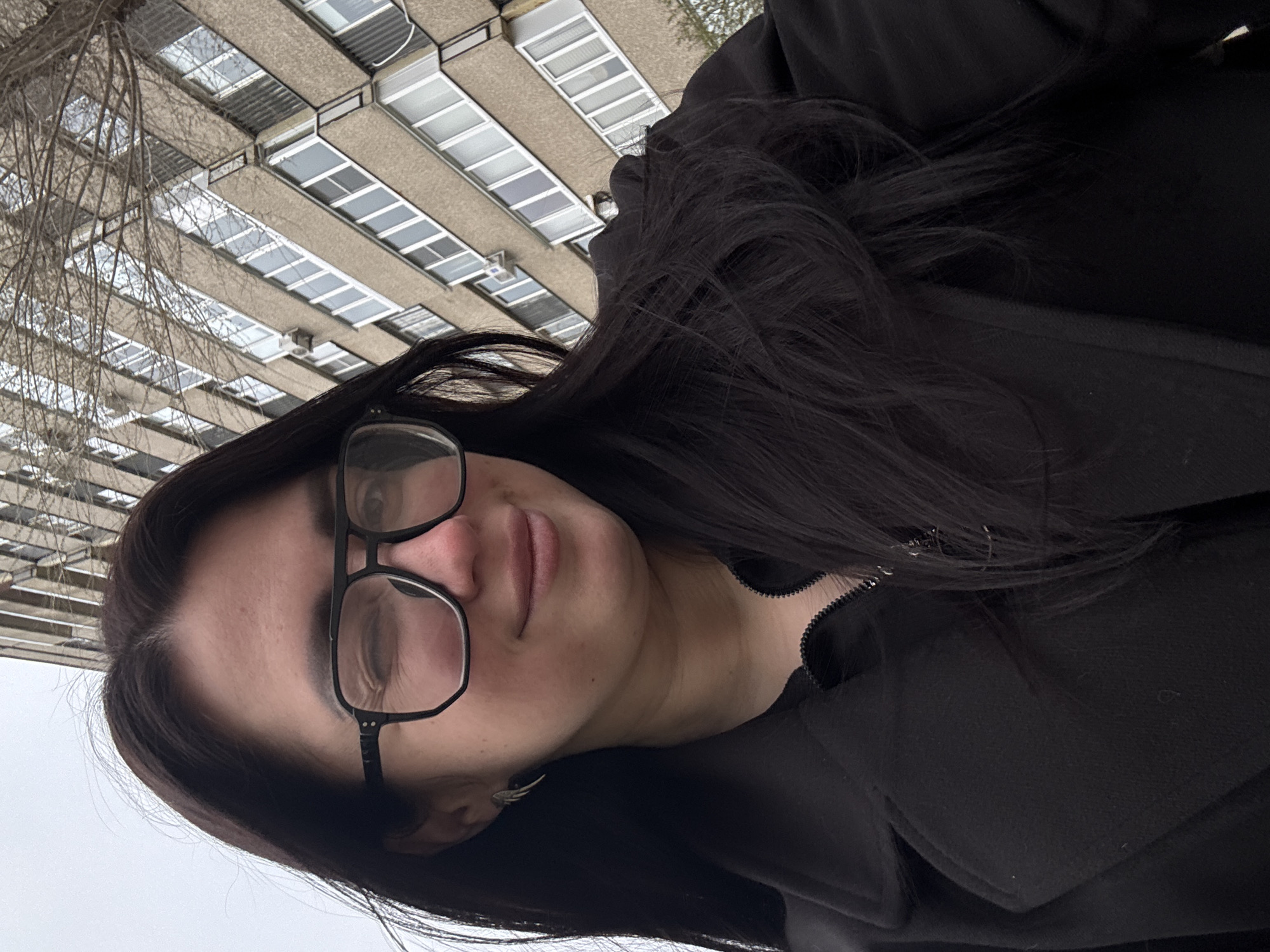 A portrait of a person with dark hair and glasses, wearing a black coat, is framed against a tall building with numerous windows and bare tree branches. The location is in the city of Unknown, Unknown.