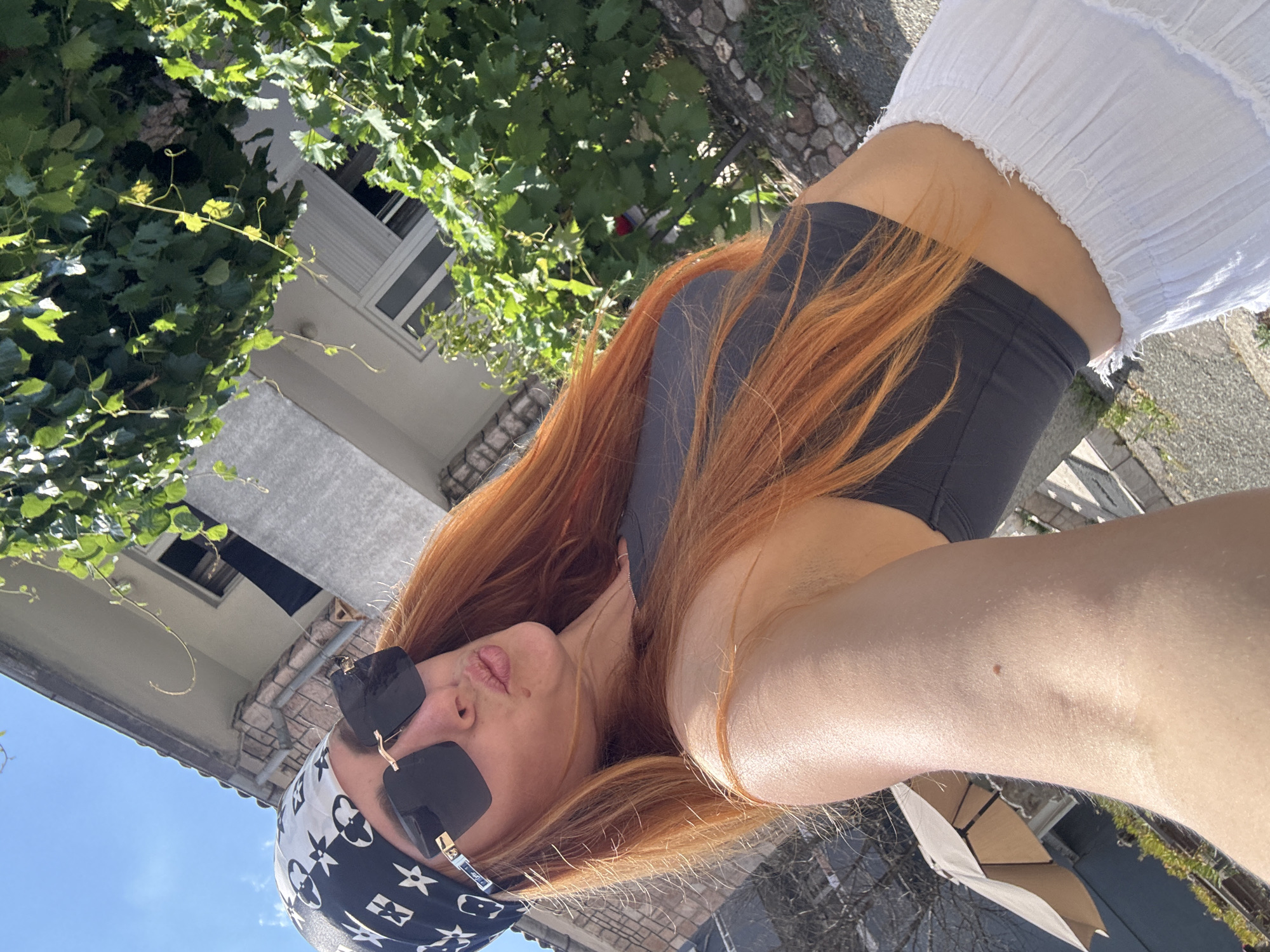 A woman with long, auburn hair is taking a selfie outdoors. She is wearing a black and white bandana with a Louis Vuitton pattern, large black sunglasses, a dark grey crop top, and a white tiered skirt. Her lips are puckered. Behind her, a stone building with windows and a white shutter is visible, partially obscured by lush green foliage. The sky is blue, and sunlight casts shadows, indicating a bright day. In the background, a partial view of a beige patio umbrella can be seen. The scene appears to be in a residential area, possibly with a garden or courtyard, consistent with a location in Budva, Montenegro.