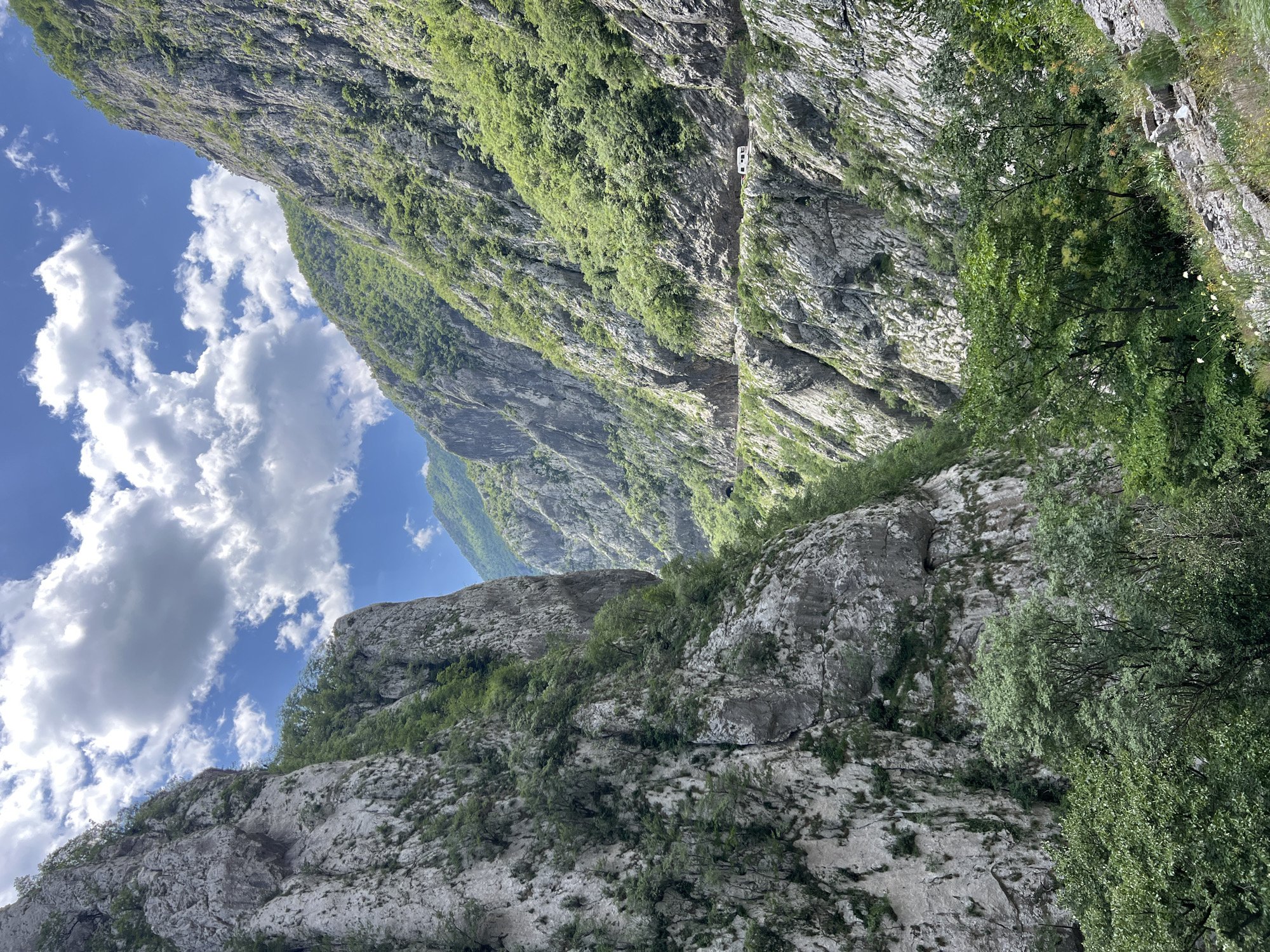 The image depicts a mountainous landscape in Vlahovići, Montenegro. The rugged terrain consists of steep cliffs and rocky outcrops covered with sparse vegetation and trees. A narrow road winds along the side of the mountain, with a white vehicle visible on it. The sky is blue with fluffy white clouds. The overall impression is of a steep, remote, and rugged environment.