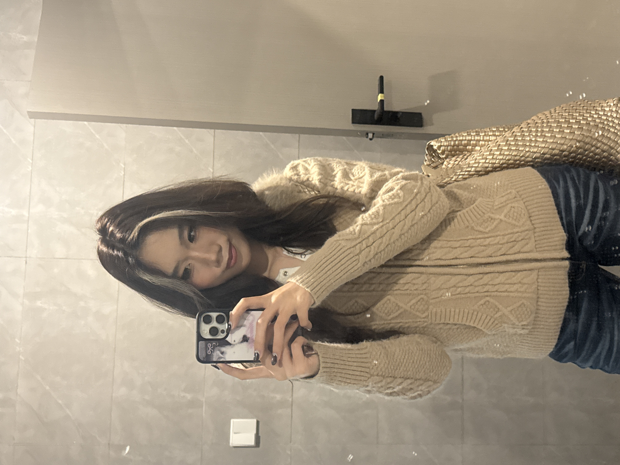 A female individual is observed taking a mirror selfie within an indoor setting in Nan'an District, China. The individual, appearing to be of East Asian descent, has long, dark hair with a blonde or lighter streak visible on the viewer's right side. She is wearing a beige cable-knit zippered cardigan over a white button-up top, paired with blue jeans. She holds a dark-cased smartphone with a triple camera system in both hands, reflecting her image. A woven golden-colored bag is visible hanging on her right side. The background comprises light-toned, rectangular wall tiles exhibiting subtle veining, with a white wall switch visible on the left. A light-colored door with a black handle, marked with a yellow band, is partially visible on the right.