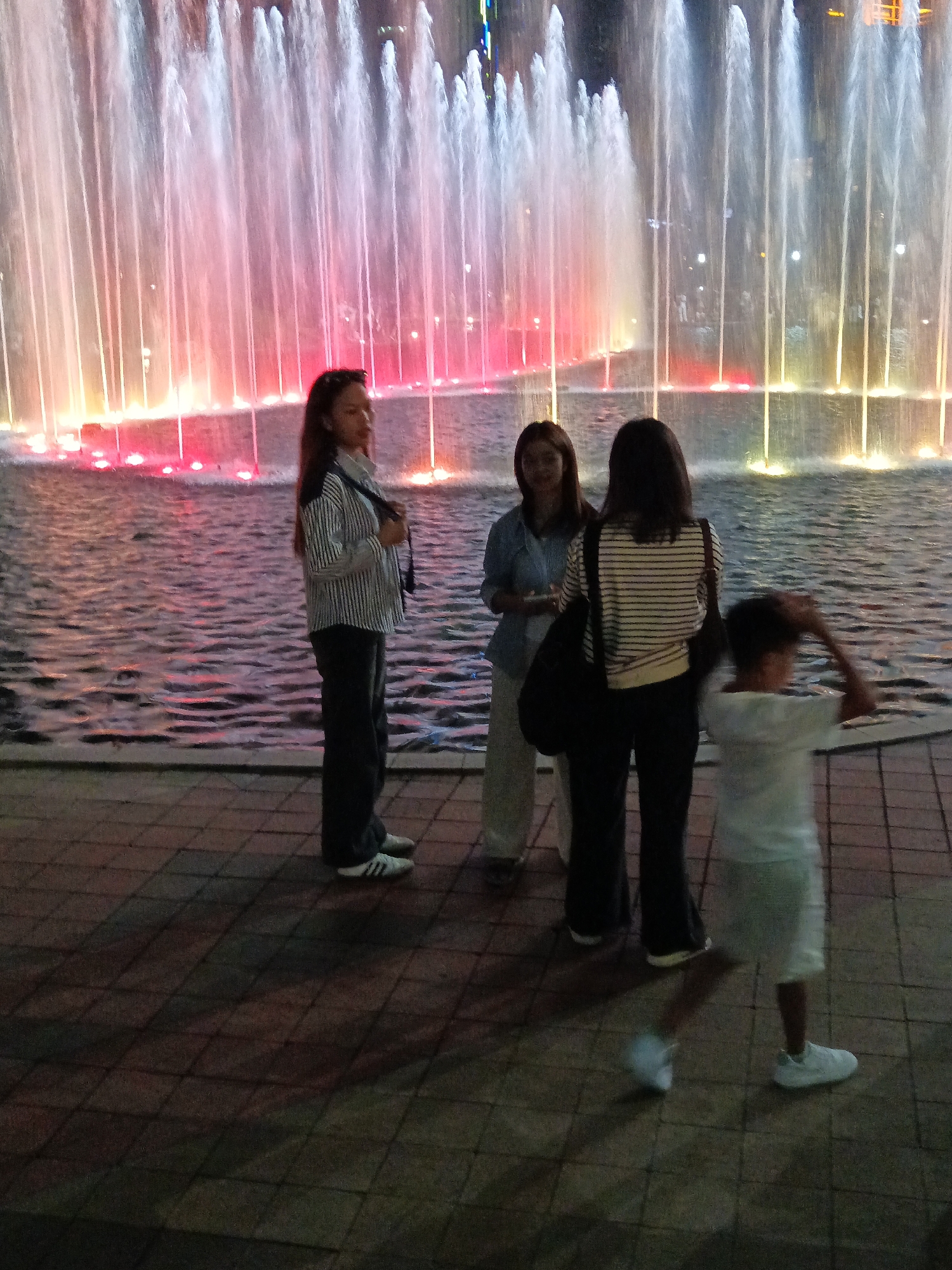 This image captures an evening scene at the Symphony Lake of the Petronas Twin Towers in Kuala Lumpur, Malaysia. A brightly lit water fountain display with multi-colored lights forms the backdrop. In the foreground, four individuals are visible. On the left, a woman in a striped shirt and dark pants stands facing left, with her right hand near her face. To her right, another woman, wearing a blue shirt and white pants, is partially visible, looking towards the fountain. Behind her, a third woman in a striped shirt and dark pants is seen from the back, holding a black bag. On the far right, a child in a white t-shirt and light-colored shorts is captured in motion, with their right arm raised. The ground is paved with patterned tiles, and the water of the lake reflects the fountain's lights.