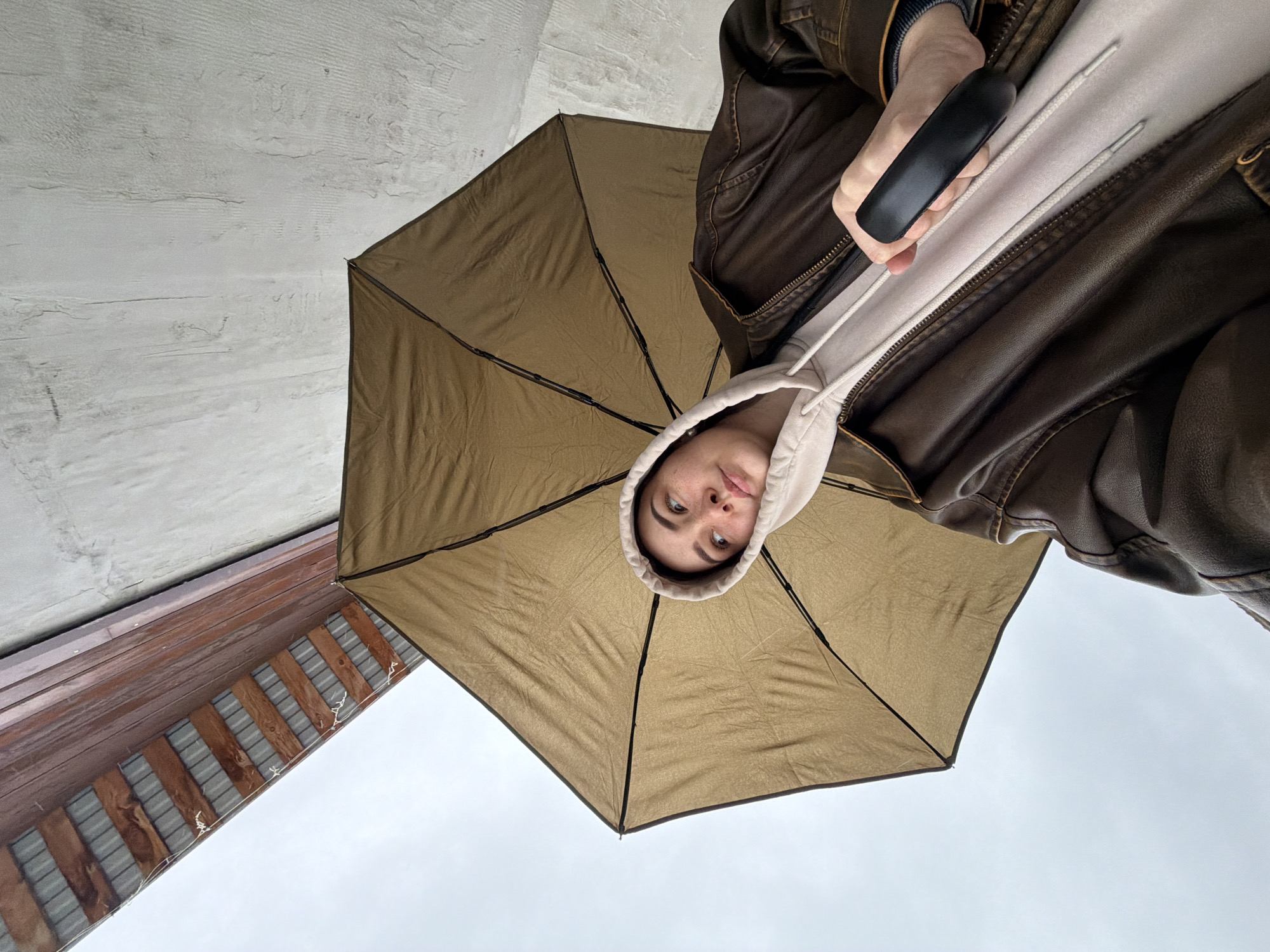 A person is holding a brown umbrella from a low angle, looking directly at the camera. The person is wearing a hooded sweatshirt and a brown leather jacket. The sky is overcast. In the background, there is a structure with wood beams and gray corrugated roofing. Above the wood structure, there is a textured white wall. There is no definitive information to prove this image was taken in Chaikovsky, Russia.