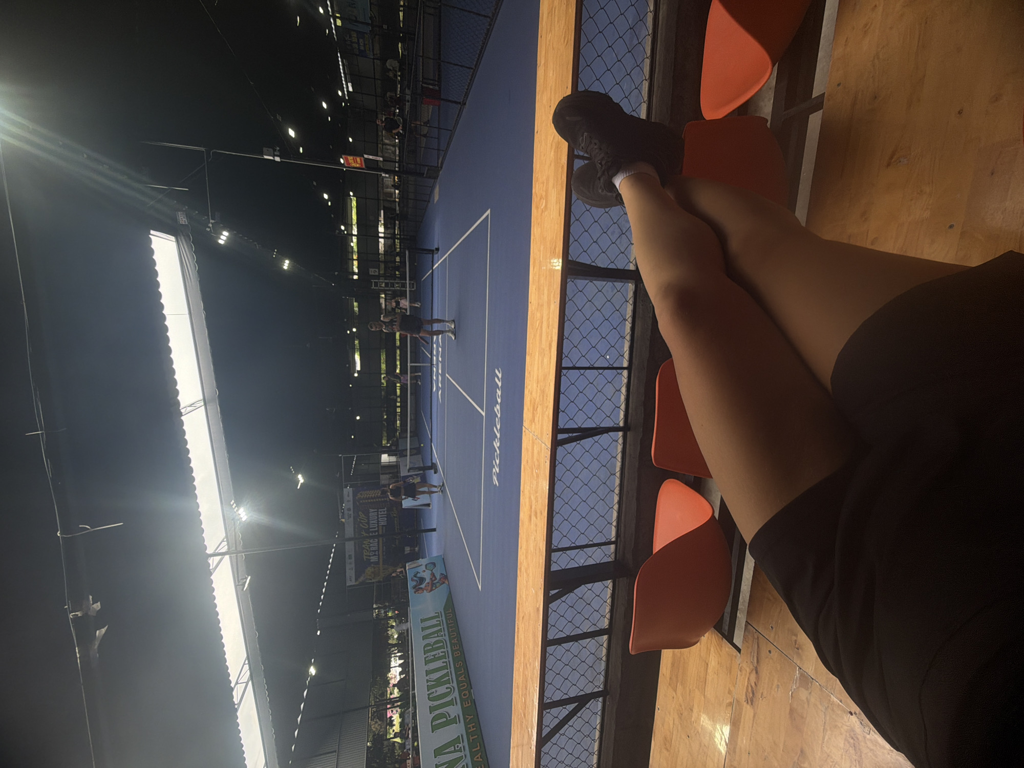 The image captures a first-person perspective from spectator seating overlooking an indoor pickleball court, rotated 90 degrees to the right. In the foreground, the legs and feet of a person are visible, clad in dark athletic shoes and socks, resting on a wooden railing or barrier. Nearby, bright orange stadium seats are arranged.
The midground features a blue pickleball court with distinct white lines and a net. Several individuals, likely players, are present on the court, engaged in or observing a game. The word "Pickleball" is clearly visible inscribed on the court surface. A chain-link fence separates the spectator area from the playing court.
In the background, an indoor arena setting is evident, characterized by a high ceiling with numerous bright overhead lights illuminating the court. Various banners are displayed, advertising events or sponsors. Text visible on one banner includes "ANA PICKLEBALL" and the slogan "HEALTHY EQUALS BEAUTIFUL." Another banner partially reads "TEAM CUP" and "M*B*G* LUXURY HOTEL," suggesting a tournament or sponsored event. The overall lighting indicates an evening or nighttime setting indoors.
The scene depicts an active sports event, specifically pickleball, taking place within an indoor facility. The context suggests this is in Nam Nha Trang Ward, Vietnam.