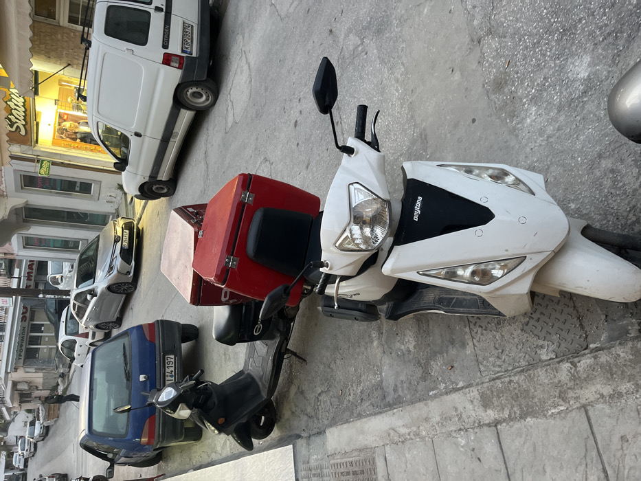 The image shows a white scooter with a red delivery box, parked on the street. The scooter is facing the camera and is situated on the left side of the image. The scooter has a black seat, black handlebars, a black rearview mirror, and a black headlight. The red delivery box is attached to the back of the scooter and has a white label on it. The street is gray and has a sidewalk on the left side. There are several cars parked on the right side of the image, including a white van with the license plate KBP 29974. The scene is set in Limenaria, Greece, at coordinates 40.627, 24.575. A person is walking on the sidewalk to the left. The sidewalk is made of gray concrete. There are two other motorcycles behind the scooter. The street has white markings. A small blue car is parked behind the white scooter. The cars are all parked on the right side of the image. There is a storefront on the right side of the image with a sign that reads "Status". There is a sign in Greek and a sign in English that reads "Cat" above the storefront. There is a building in the background. There are trees and a small white car in the distance. The image was taken during the daytime, but the lighting is somewhat dim. The sky is overcast.
