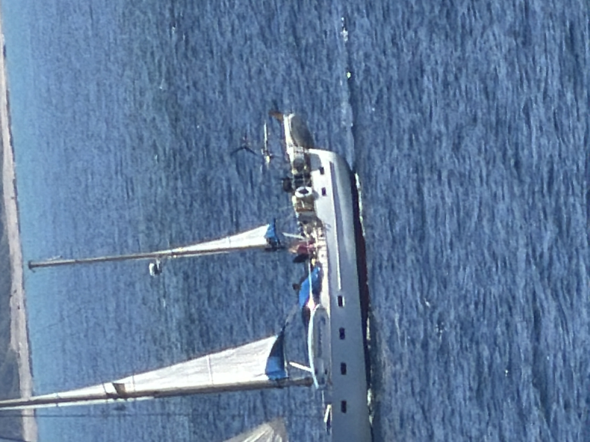 In the Municipal Unit of Kastro - Kyllini, Greece, a white sailboat with blue accents on its sails navigates the blue waters of the Ionian Sea. The sailboat has two masts and appears to be a monohull design. A few people are visible on the deck of the boat. The water is slightly choppy, with the distant shore visible in the upper portion of the image. The scene is illuminated by natural daylight.