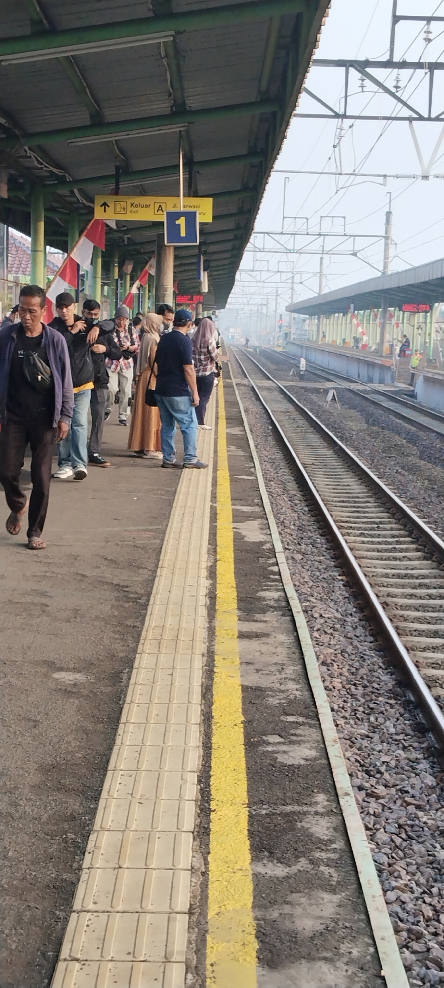 The image depicts a train station platform in Daru, Indonesia, with multiple individuals waiting. The platform is covered by a green-painted roof supported by pillars. Yellow and white tactile paving runs along the edge of the platform, bordering the railway tracks. Several people are visible standing on the platform, some are walking, and one individual in the foreground is walking away from the camera. A yellow sign indicates "Keluak A Exit" and the street "Jl. Arwani". A blue sign with the number "1" is also present. Red and white Indonesian flags are displayed on poles along the platform. The railway tracks are to the right of the platform, and a second platform can be seen across the tracks, also with people and a covered roof. The sky appears hazy.