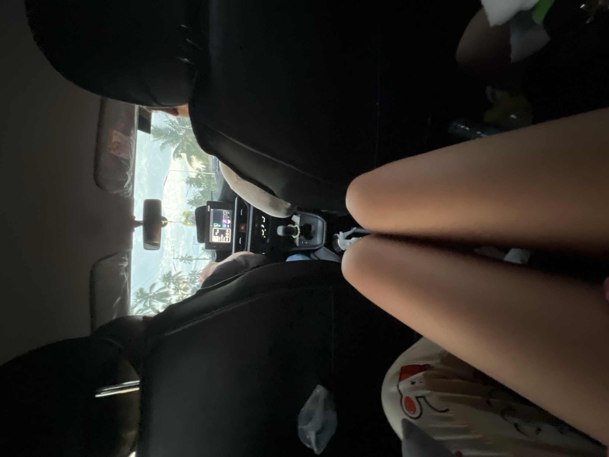 A view from the back seat of a car, likely taken by a passenger in Davao City, Philippines, showing a partial view through the front windshield. Palm trees and light-colored buildings are visible in the distance. The driver is present, with the headrest of the driver's seat visible. Also visible are the driver's dashboard and a small navigation screen. In the back seat, the leg of a passenger is present, and they appear to be holding a plate with some food on it.