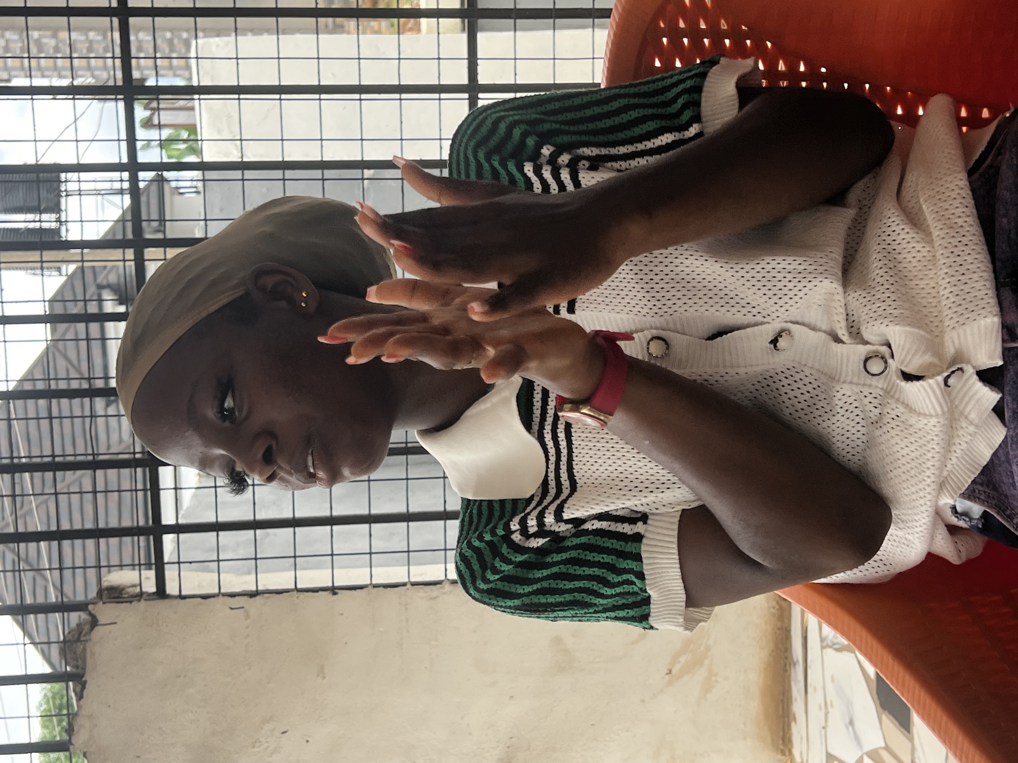 A young Black woman with dark skin and dark hair, wearing a light brown headscarf, a white polo shirt with green and black striped sleeves, and a red wristband, sits in an orange plastic chair. She has her hands clasped in front of her, and her mouth is slightly open as if she is speaking or singing. The woman is positioned in front of a white, rough-textured wall and a metal grate that obstructs the view of the background. Through the grate, a glimpse of buildings and the sky can be seen. The woman's pose and expression suggest she is engaged in an activity, possibly a performance or a conversation. The setting appears to be outdoors, possibly in a courtyard or patio area, given the open view through the grate and the presence of the plastic chair. The environment seems to be in Nkpor, Nigeria, characterized by the architectural style and the lighting conditions.