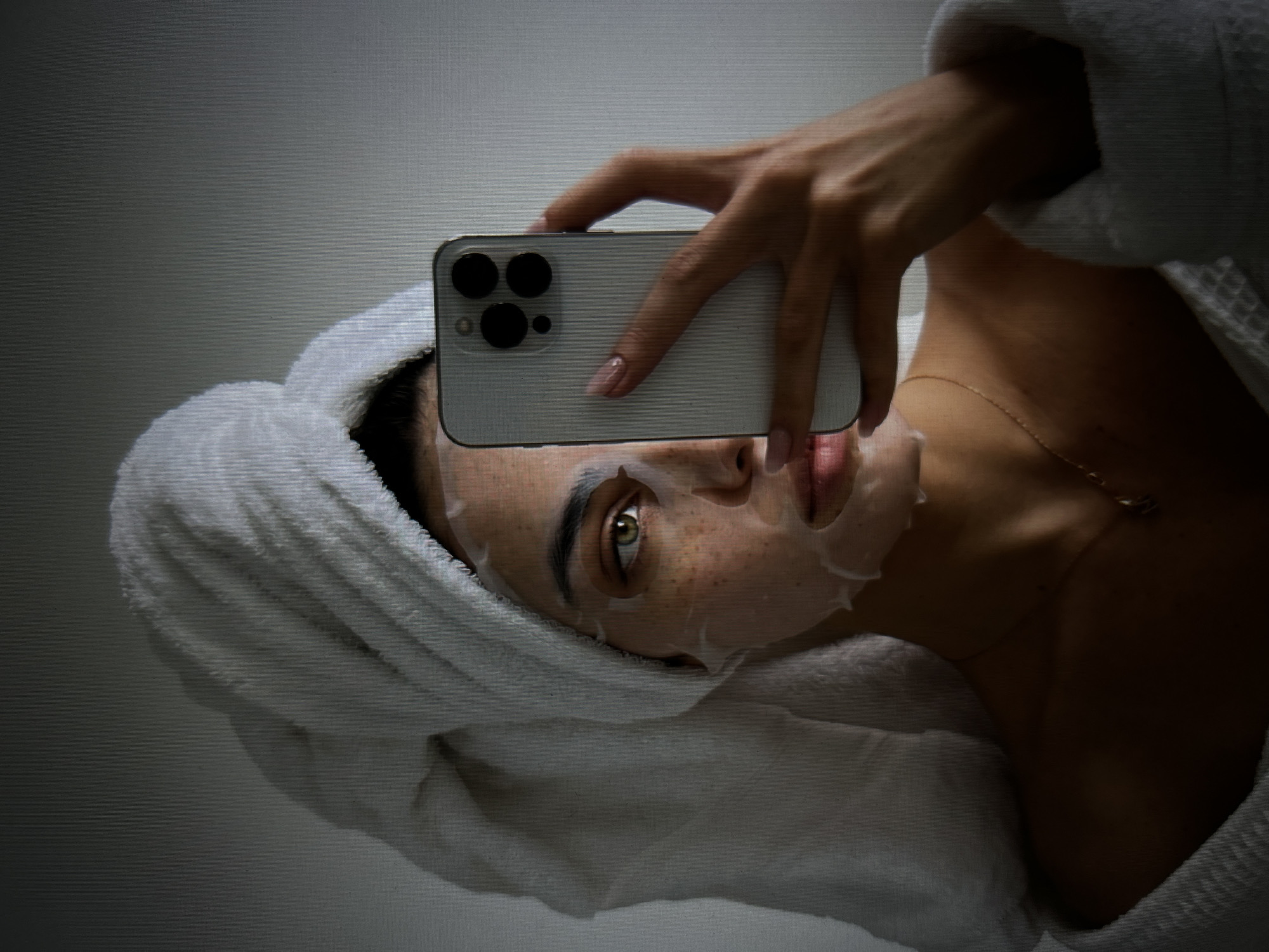 A young woman with a towel wrapped around her head and a sheet mask on her face is taking a selfie with a smartphone. She is wearing a white bathrobe. Her right hand holds the phone, and her left hand is not visible. The background is a plain, light-colored wall. The image is shot from a medium-low angle.