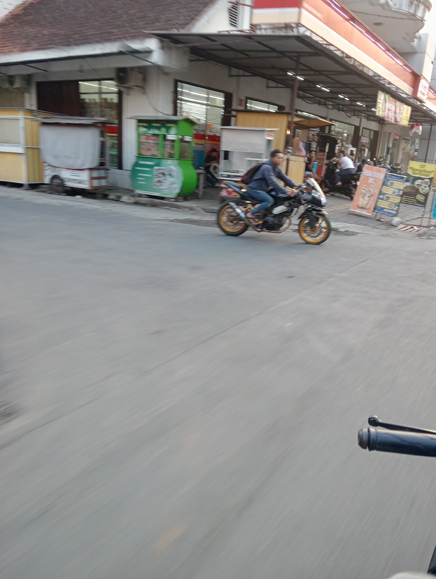 The image depicts a street scene in Grabag, Indonesia, at what appears to be late afternoon or early evening. The foreground is dominated by a blurred, grey asphalt road, suggesting motion, likely from a vehicle. Partially visible in the lower right corner is the handlebar and brake lever of a motorcycle, indicating the viewpoint of the photograph.
In the middle ground, a person is riding a black and white motorcycle with gold-colored rims, moving from left to right. The rider is wearing a blue jacket and blue jeans, with a dark backpack slung over their shoulder. The motorcycle itself has a custom exhaust.
The background features a row of buildings with awnings. To the left, there are at least two food stalls or small kiosks. One is yellow and white with a white cart, and another is a prominent green structure with text and images visible. Further to the right, the buildings appear to be more permanent structures, possibly shops or a convenience store, with signage visible. Several other motorcycles are parked in front of these buildings, and some people can be seen milling about. Yellow and blue advertising posters are displayed on stands in front of the buildings. The overall impression is a busy commercial area with active street life.