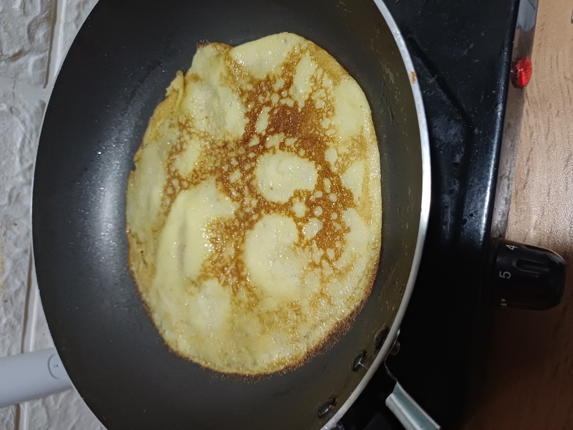 A close-up, high-angle shot shows a single, golden-brown crepe cooking in a black non-stick frying pan. The crepe has a slightly uneven, circular shape, with some areas appearing lighter yellow and others a deeper, toasted brown, indicating it is in the process of being cooked. The pan sits on a black stovetop. In the background, a white tiled wall is visible, with a small portion of a white appliance on the left. A black control knob with white markings, including the number "5", is present on the stovetop. The scene is domestic and appears to be in a kitchen. There is no information to suggest this is in Sochi, Russia.