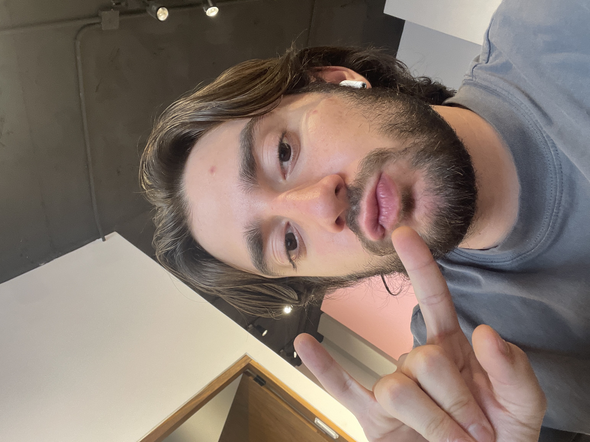 A medium close-up, slightly rotated portrait depicts a light-skinned man with long brown hair and a full dark beard. He is wearing a gray t-shirt and has a white wireless earbud in his ear. He is holding his hand up, forming a "V" sign with his index and middle fingers.
The background consists of a grey ceiling with track lighting and a white and pink wall. A wooden door frame is visible in the lower left corner. Without more information, it is not possible to confirm the location in Mexico City, Mexico.