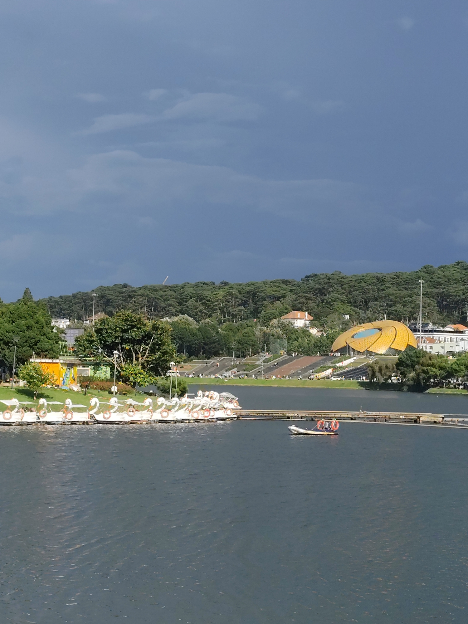 The image depicts Xuan Huong Lake in Da Lat, Vietnam, under a dramatic, overcast sky. In the foreground, a large body of dark, rippling water dominates the lower half of the frame. Along the left bank, a line of white swan-shaped paddle boats is moored. These boats are equipped with orange life preservers. In the middle ground, a narrow wooden pier extends into the lake, and a small motorboat with two people wearing orange life preservers is passing by.
Beyond the pier, the landscape rises into a densely forested hillside. On the hillside, several buildings are visible, including a distinctive golden, petal-shaped structure that appears to be a modern pavilion or amphitheater. The trees are predominantly dark green pines. The sky is a deep, brooding gray with some lighter patches, suggesting impending rain or recent downpours. The overall atmosphere is serene yet somber.