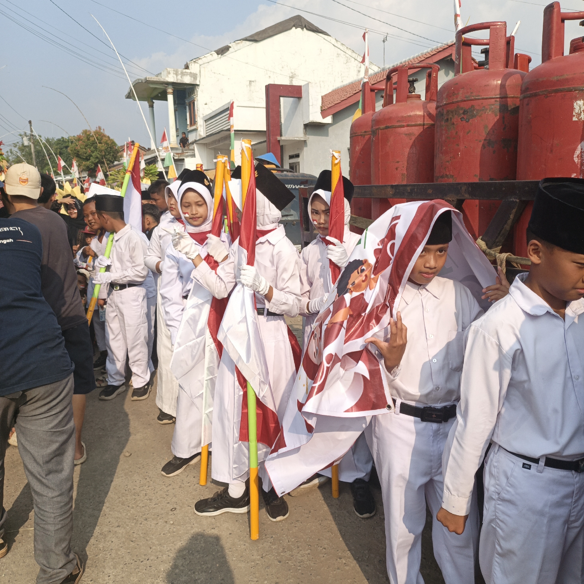 In Tanahbaya, Indonesia, a group of schoolchildren, dressed in white uniforms and black caps, participate in a parade. Several students carry flags and banners. One student is covered by a large banner depicting a cartoon figure. In the background, large red gas cylinders are visible, along with houses and power lines. The sky is clear and the sun is shining.