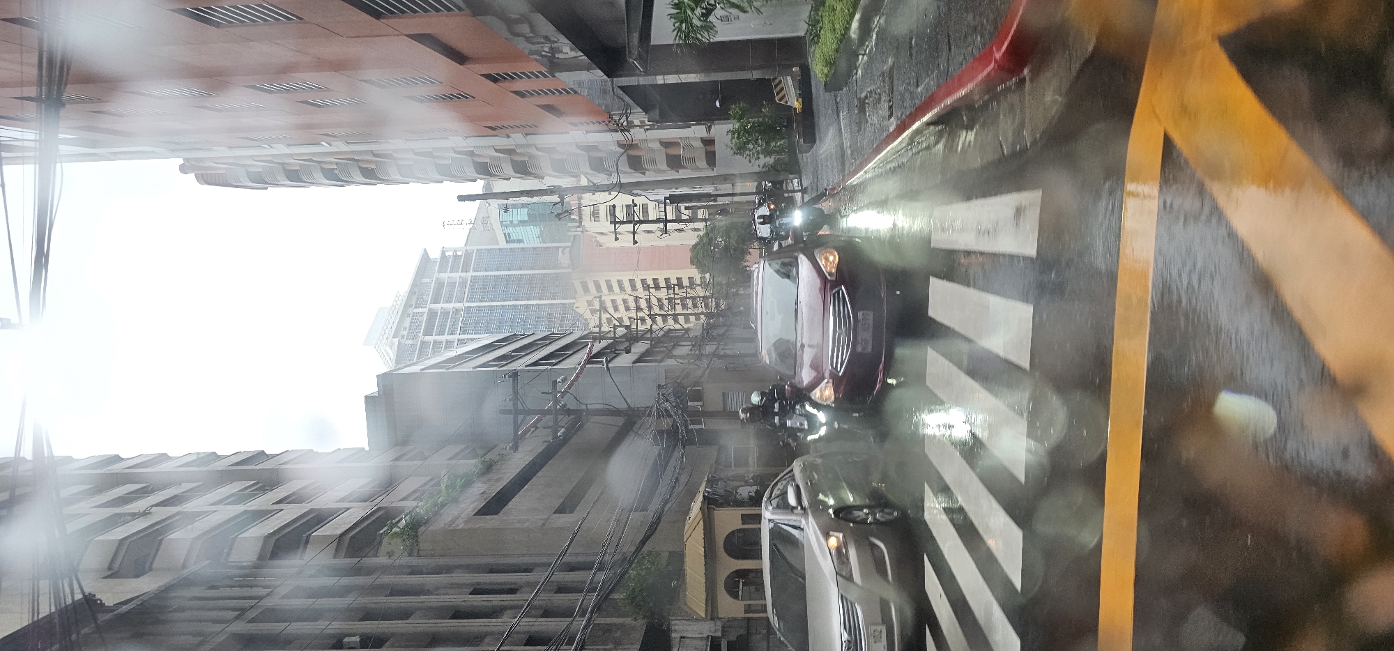 This is a mid-shot, vertical-oriented, photograph taken from inside a vehicle, looking forward through a wet windshield. The scene captures a street in Makati City, Philippines, during a rainy day. The road is wet, with visible reflections and streaks from water droplets on the glass.
In the foreground, a pedestrian crosswalk with white stripes is clearly visible on the wet asphalt. A yellow traffic line is also present.
In the middle ground, several vehicles are present. A silver sedan is stopped on the left side of the crosswalk, facing forward. To its right and partially on the crosswalk, a maroon sedan is also stopped, facing forward. Between these two cars, a motorcyclist wearing a helmet and dark clothing is stopped on the motorcycle, facing forward. There are other vehicles and motorcyclists further down the street, some of which are difficult to discern due to the rain and distance.
The background is dominated by tall buildings. On the left, a concrete building with visible balconies and windows rises upwards. To its right, a modern, multi-story building with a glass facade is prominent. On the right side of the image, another tall building with a reddish-orange facade and rows of windows is visible. Utility poles with numerous wires are scattered throughout the scene, adding to the urban environment. The sky is overcast and bright, suggesting a daytime scene under heavy rain. The overall impression is a typical urban street scene in the Philippines during a downpour.
