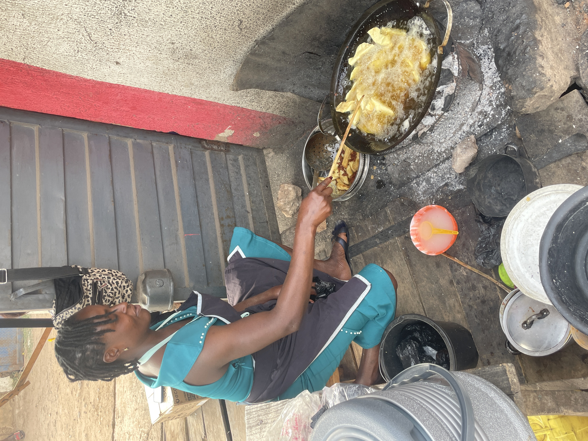 This outdoor image, captured in Jos, Nigeria, depicts a woman deep-frying food over an open fire under bright daylight, suggesting a street food stall or an informal cooking setup.
The woman, likely of African descent, has dark skin and dreadlocked hair. She is dressed in a teal top with white dotted embellishments and dark trousers with matching teal and white accents. She appears to be seated on wooden planks, focused intently on the cooking process. Behind her, a leopard-print bag is visible, along with some stacked boxes, one of which partially displays the text "ECO" and another "LE FOUR".
In front of her, a large, dark metal pan is filled with bubbling oil, in which numerous pieces of food, likely plantains or yams, are frying to a golden brown. She uses a long wooden stick to stir and manage the food. Beside the frying pan, a metal bowl contains a batch of already fried food and a slotted metal skimmer. The pan rests on a rudimentary stove setup, indicated by ash and a collection of stones or bricks that form a hearth for an open fire.
The surrounding area features a dark gray corrugated metal gate or wall on the left, topped by a textured light-colored wall with a prominent horizontal red stripe. The ground is a mix of dirt and rough surfaces. Various utilitarian items are scattered around the cooking area, including several dark buckets (some containing plastic bags), other metal pots with lids, and a bright orange bowl with a yellow spoon. The scene portrays industrious activity in an everyday, informal outdoor environment.