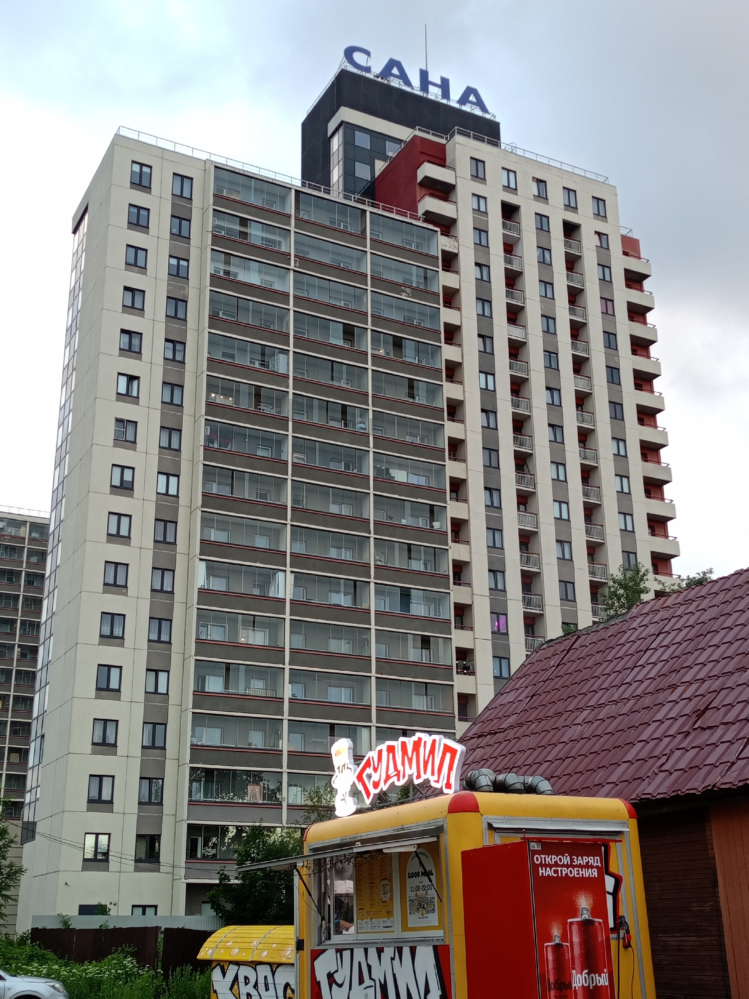 In an urban exterior scene, a tall, modern apartment building with numerous windows and balconies dominates the background. The building features a cream-colored facade with gray vertical accents. On top of the building, large blue letters spell out "САНА" (SANA). In the foreground, a yellow food truck is parked, with a red section on its side displaying text and images, including two red cans labeled "Добрый" (Dobryi). The food truck also has a red illuminated sign with white Cyrillic text that appears to read "БУДМИЛ" (BUDMIL). A portion of a red-tiled roof of a smaller building is visible in the lower right quadrant of the image, and some greenery is present in the lower left. The sky is overcast.