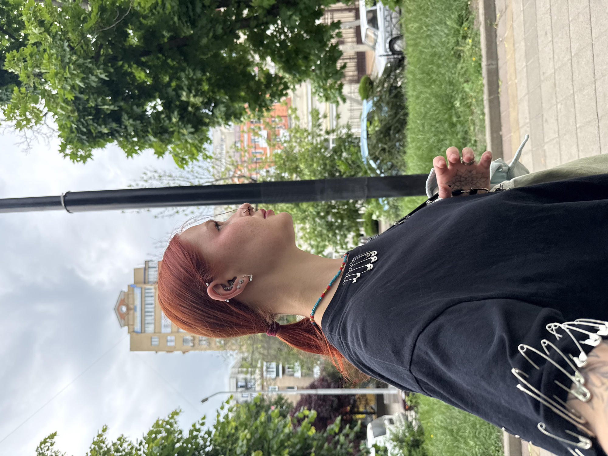 A young person with red hair is looking up and to the left in an outdoor urban environment. They are wearing a black t-shirt decorated with safety pins along the sleeve and a colorful beaded necklace. The person has multiple ear piercings. Part of their hand is visible, revealing a tattoo. The background includes trees, a sidewalk, a building, and a street light. Based on architectural styles and the general environment, the location is possibly in the city of Rostov-on-Don, Russia.