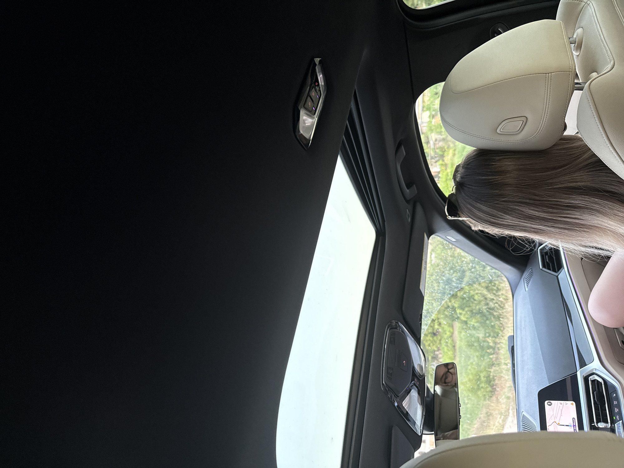 The image is taken from inside a car, looking forward and slightly upwards through the sunroof. The sunroof is open, revealing a bright sky and green foliage outside. The interior of the car is visible, including the black headliner, a rear-view mirror reflecting a person wearing glasses, and the dashboard with a navigation screen showing a map. Two passengers are visible: one in the front passenger seat (partially visible) and another in the rear, leaning their head against the headrest, their long blonde hair falling over their shoulder. The exterior environment appears to be rural or semi-rural, with trees and vegetation visible through the open sunroof and windshield. The image does not contain any information that allows for identification of the specific location within the city of Sărata-Mereșeni, Moldova.