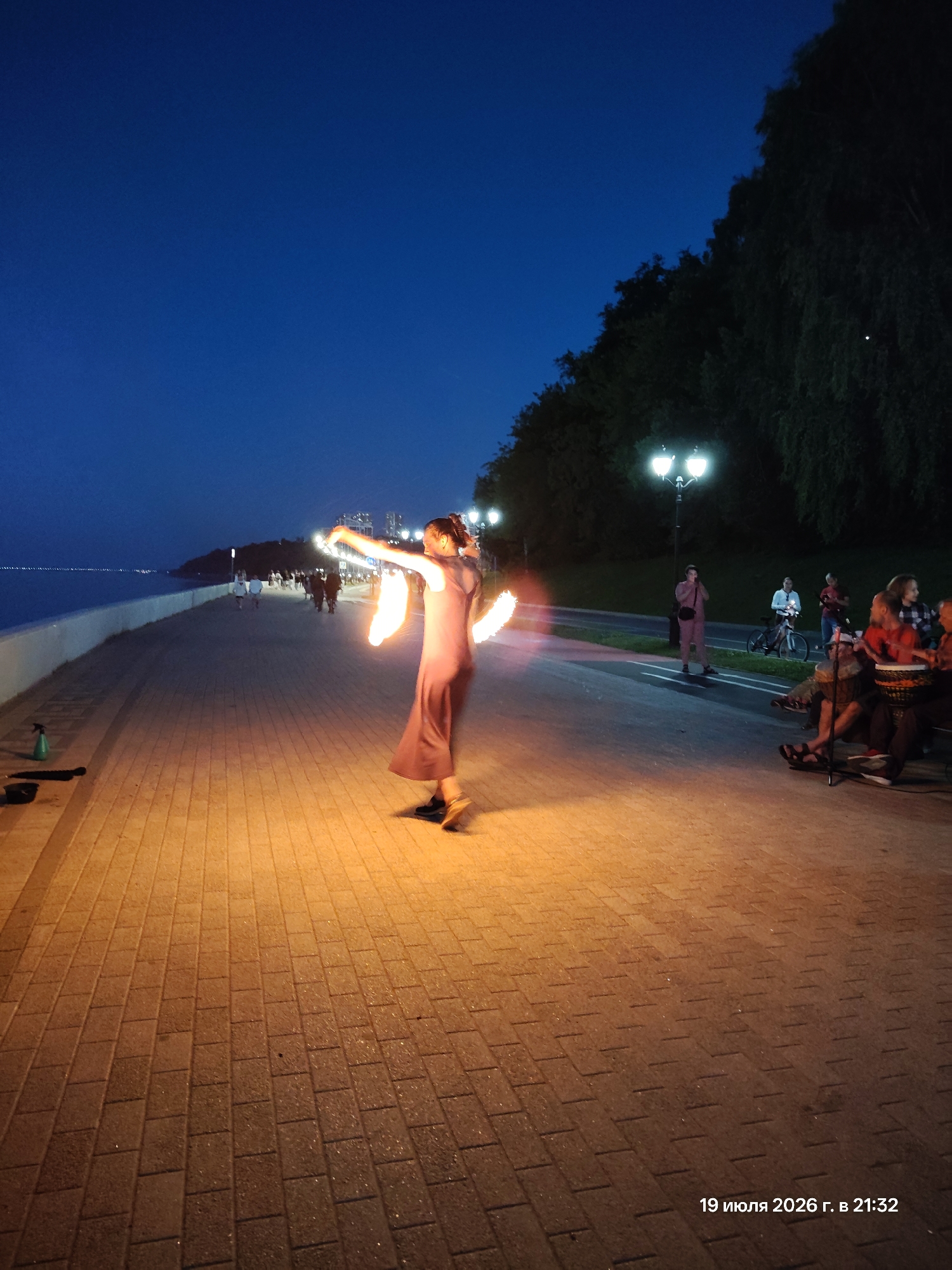 A wide shot shows a waterfront promenade at dusk. In the foreground, a woman in a dark, flowing dress performs with fire, her arms extended to create glowing rings of light. The promenade is paved with reddish-brown bricks, illuminated by the warm glow of the fire and streetlights. To the left, a green spray bottle and black object rest on the ground. Further down the promenade, a crowd of people walks and stands, their silhouettes dark against the twilight sky. In the background, trees line the edge of the promenade, their dark foliage contrasting with the illuminated path. The water of a large body of water stretches out to the left, meeting the horizon. In the middle ground, to the right, a group of people is gathered, some sitting and appearing to play drums. The sky is a deep blue, transitioning to a lighter shade near the horizon. In the far background, city lights are visible. The date and time "19 июля 2026 г. в 21:32" are visible in the lower right corner of the image.