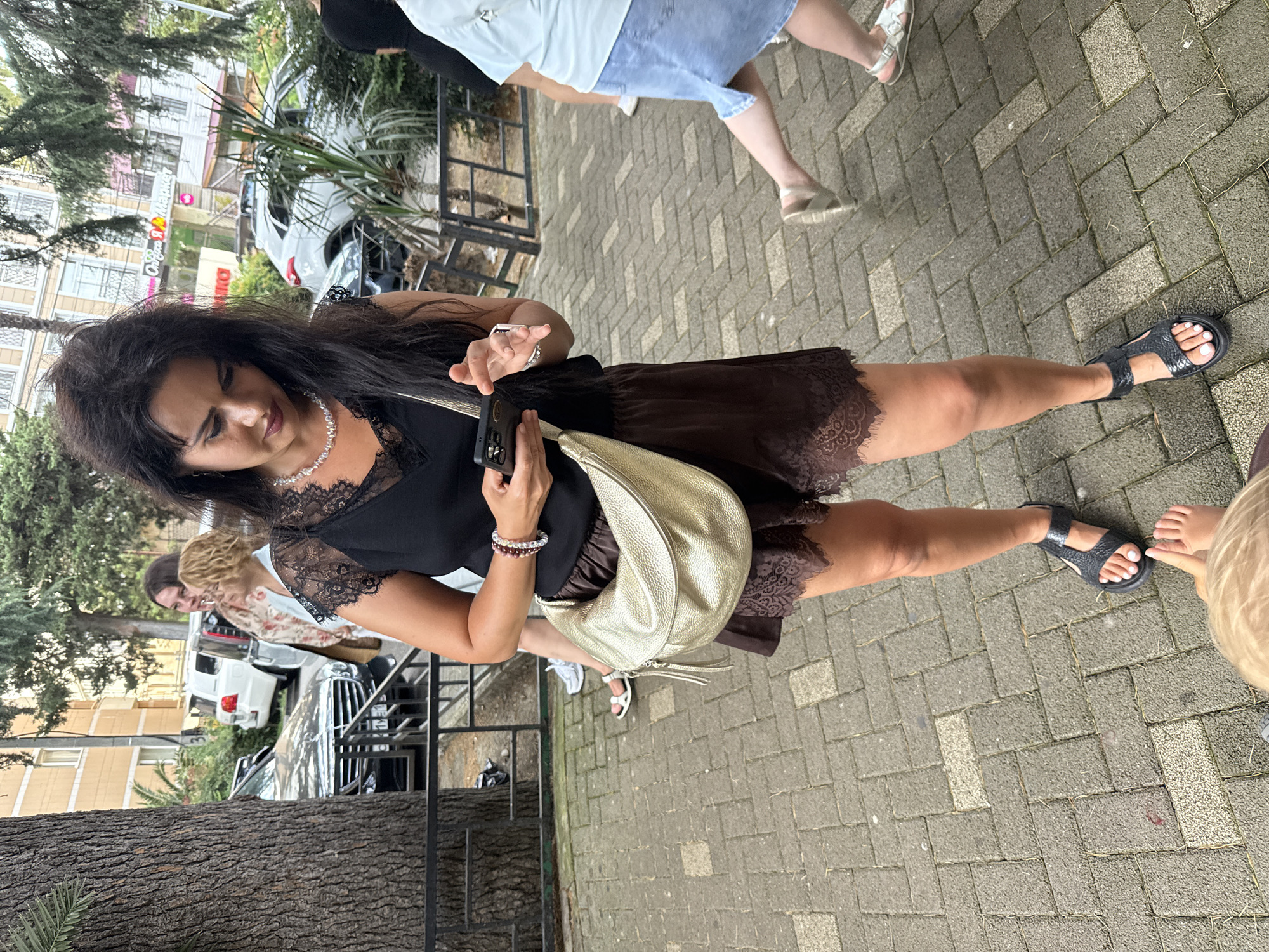 The image shows a woman with dark, voluminous hair, wearing a black lace-trimmed top and a dark brown skirt. She is holding a smartphone in her right hand and has a metallic gold purse slung over her shoulder. She is wearing beaded bracelets on her left wrist and dark sandals. To her left, a child with blonde hair is visible, reaching out towards her. Further in the background, other individuals are partially visible, one wearing a light blue top and denim skirt, and another in a patterned shirt.
The scene appears to be outdoors, on a paved walkway made of interlocking bricks. In the background, there are buildings with pale yellow and white facades, some with signage that is partially obscured. A white vehicle is parked behind a metal fence. The presence of trees and the general atmosphere suggest a daytime setting, possibly a warm day, given the light clothing worn by some individuals. The woman's expression is somewhat averted, looking down at her phone. The overall context suggests a casual outing or stroll in an urban or semi-urban environment, possibly a resort town like Sochi, Russia, as indicated by the location context.