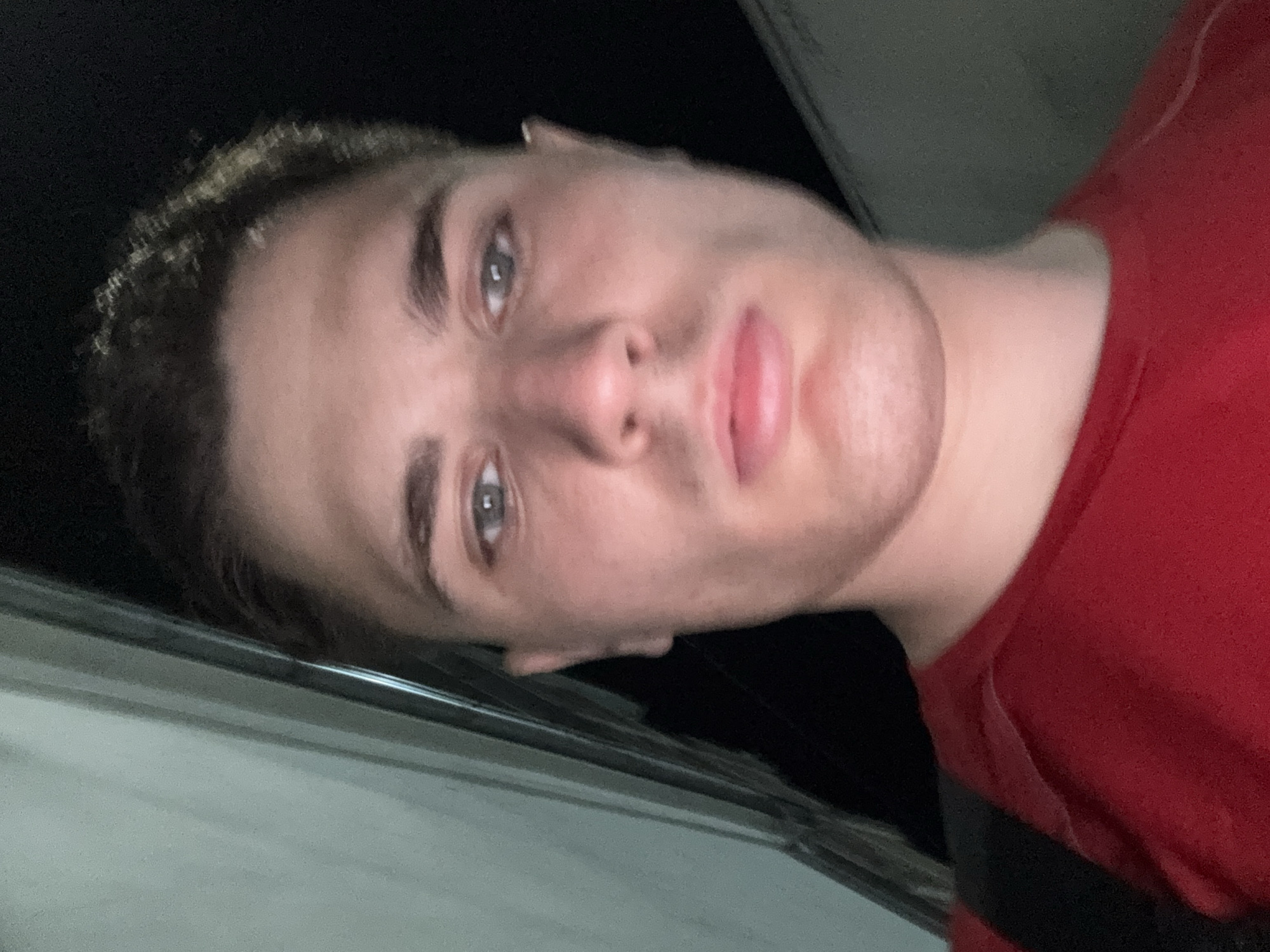 A young Caucasian male with short brown hair and blue eyes is pictured in a close-up selfie. He is wearing a red t-shirt and a black backpack strap is visible. The background is dark, with a blurred light-colored vertical structure on the left side of the frame, possibly a building facade or a door frame. The image appears to have been taken at night, as indicated by the dark background and the lighting on the subject's face. The setting does not provide specific clues to identify a location within Stara Zagora, Bulgaria.