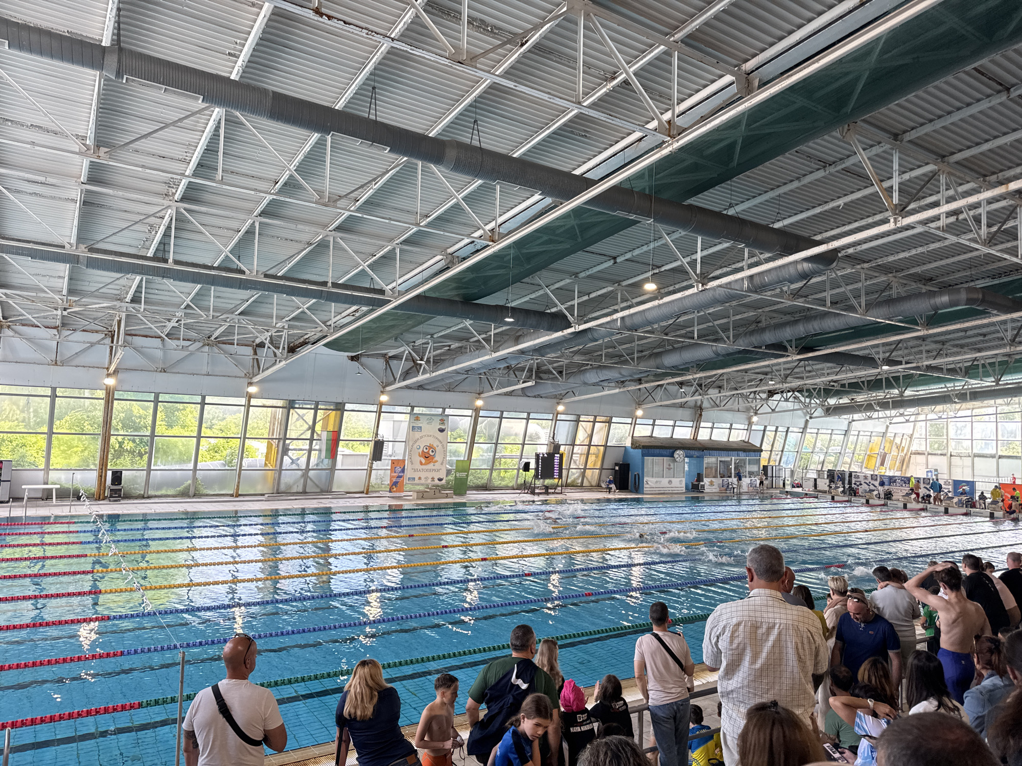 The image shows an indoor swimming pool with several lanes marked by lane ropes. Swimmers are in the water, participating in what appears to be a competition. Spectators are observing from the side of the pool. The location appears to be the "Slatina" swimming complex in Sofia, Bulgaria, given the prominent signage visible. The building has a high ceiling with exposed metal beams and industrial-style air ducts. Large windows provide natural light.