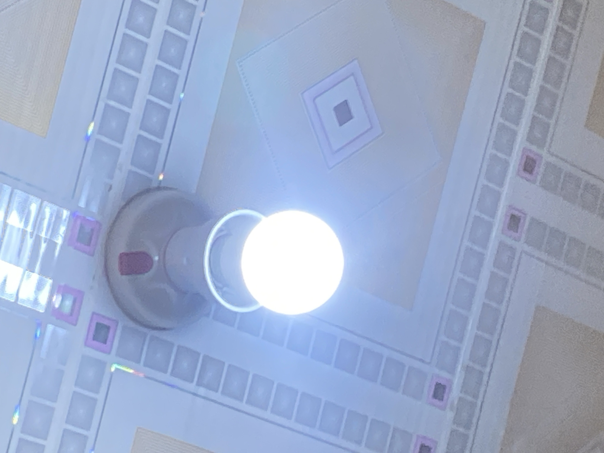 A bright, round LED lightbulb is illuminated, hanging from a simple fixture. The lightbulb is the central subject and is positioned against a backdrop of a ceiling with a geometric pattern. The ceiling appears to be made of square tiles, some of which have subtle pink or purple square inlays. There are also larger, diamond-shaped patterns on the ceiling, with layered square outlines. The lighting is bright and cool-toned. The image does not contain any people or actions, and the environment appears to be indoors, likely a room with a decorative ceiling. The location in Jauro Yinu, Nigeria, is not visually discernible from the image itself.