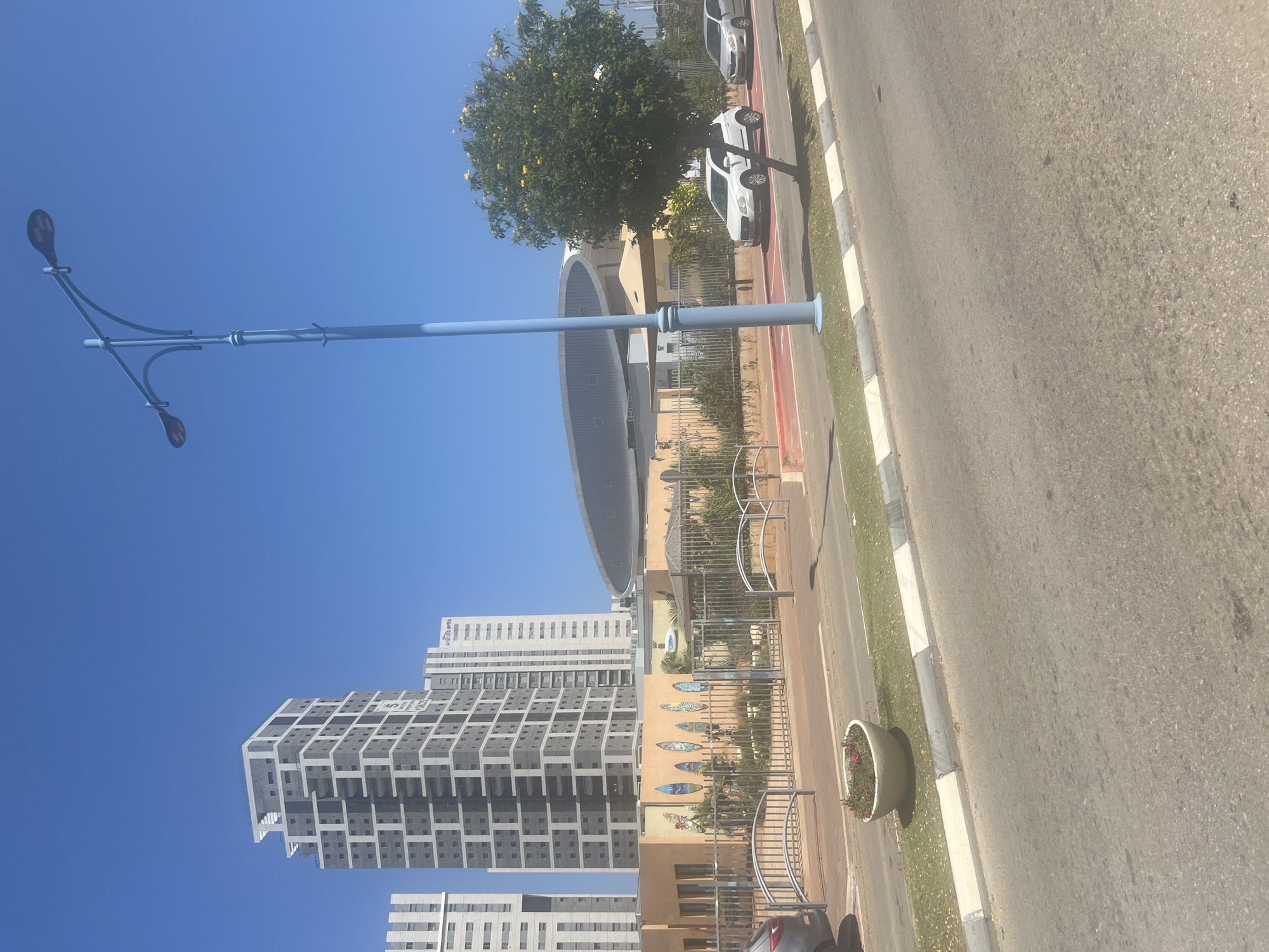 The image depicts a street scene in the city of Hadera, Israel, on a clear, sunny day. In the foreground, a paved road with a grass median strip bordered by a concrete curb with grey and white markings runs across the bottom of the frame. A large, white, oval-shaped planter with pink flowers is situated on the grass median.
Dominating the middle ground is a low-rise, light-orange building with several windows. It is surrounded by a metal fence. The building's facade is decorated with a series of colorful, surfboard-like shapes. Behind this building, a large, grey, oval-shaped structure with a metallic sheen is visible, partially obscured by a leafy green tree with yellow flowers.
To the left of the orange building, two tall, modern apartment buildings with grey and white facades stand against the bright blue sky. The building on the left is predominantly grey with white horizontal and vertical accents, showcasing a stepped architectural design. The building to its right is primarily white with grey sections and some visible signage.
A light blue lamppost with two curved arms, each holding a street lamp, stands prominently on the right side of the image, extending into the blue sky. In front of the orange building, two cars are parked on a red-marked lane, a white compact car and a silver sedan. A grey SUV is partially visible on the far left of the image, parked on the road. The overall environment suggests a well-maintained urban area with residential and potentially commercial or educational facilities.