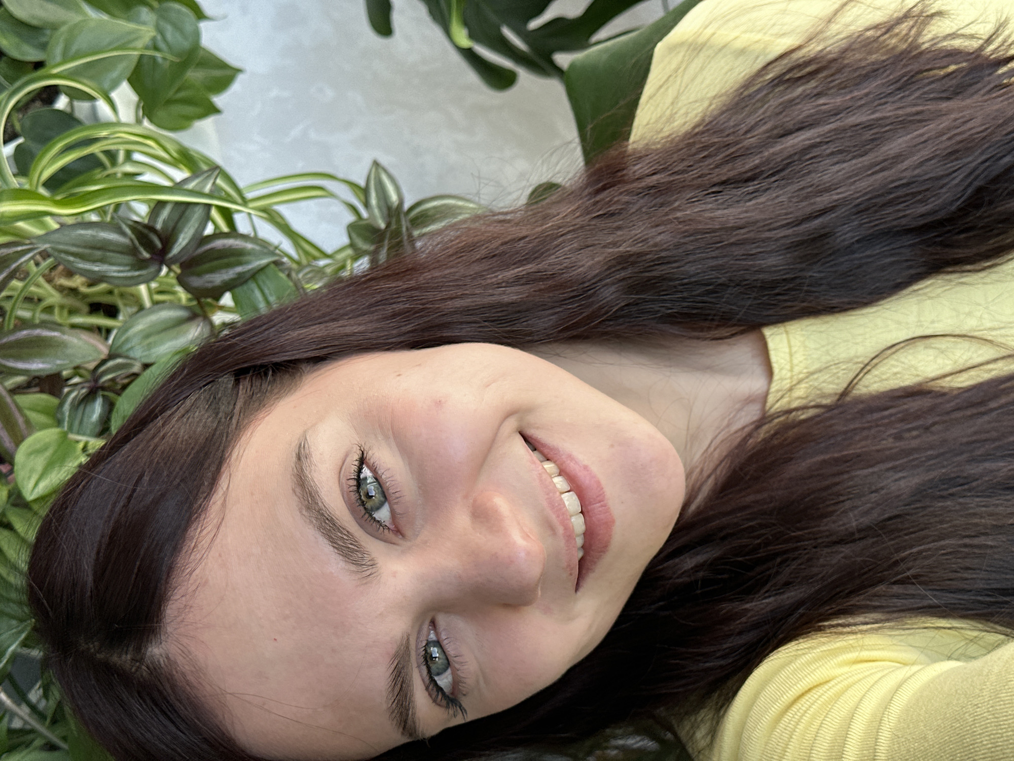 A selfie shot captures a young woman with long, dark brown hair, wearing a yellow shirt. She is smiling and looking directly at the camera, which is positioned at an angle looking up at her face. Her eyes are a light green or hazel color. To the left of the woman, various houseplants are visible with green and purple leaves. In the background, additional greenery can be seen, suggesting the photo was taken indoors, possibly in a home or a location with potted plants. There is no evidence to suggest the location is Chaikovsky, Russia.