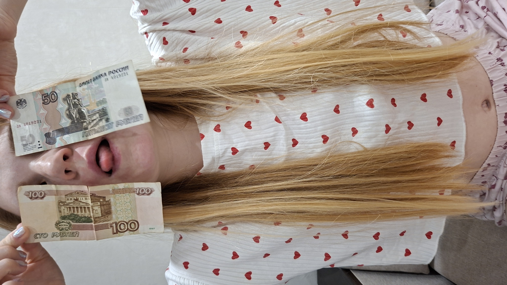 The image shows a light-skinned individual lying on their back, possibly on a sofa. They have long, blonde hair spread out around them. The person is wearing a white top with small red heart patterns. A portion of their torso and abdomen is visible, including a navel.
The person is holding two Russian Ruble banknotes. A 50 Ruble note is held over one eye, and a 100 Ruble note is held over the mouth/chin area. The person's mouth is open and their tongue is visible.
The 100 Ruble note depicts the Bolshoi Theatre in Moscow, while the 50 Ruble note features a statue of a woman seated, along with a representation of Saint Petersburg. The location mentioned in the request, Chaikovsky, Russia, is not visible in the image and doesn't seem to be directly related to the banknotes or the person depicted. The background is a plain, light-colored wall.