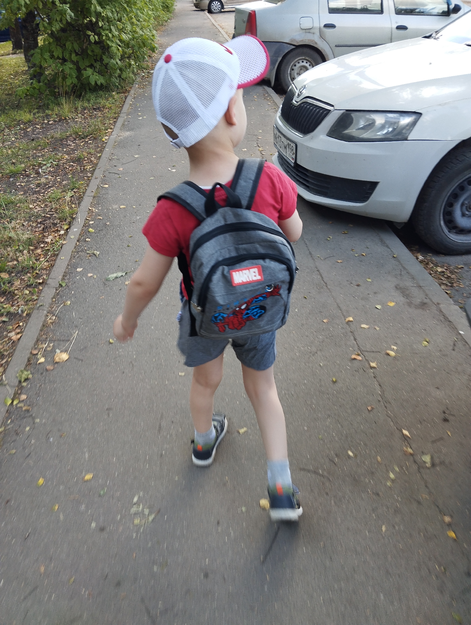 A young child wearing a white baseball cap with a red brim and a Marvel backpack walks on a paved sidewalk next to a street. The child is facing away from the camera and is wearing a red t-shirt and grey shorts. Two cars are parked on the street to the right of the child, one white and one silver. The sidewalk is lined with green bushes and trees. The ground is littered with fallen leaves, suggesting it is autumn. This scene is consistent with a residential area in the city of Kingisepp, Russia.