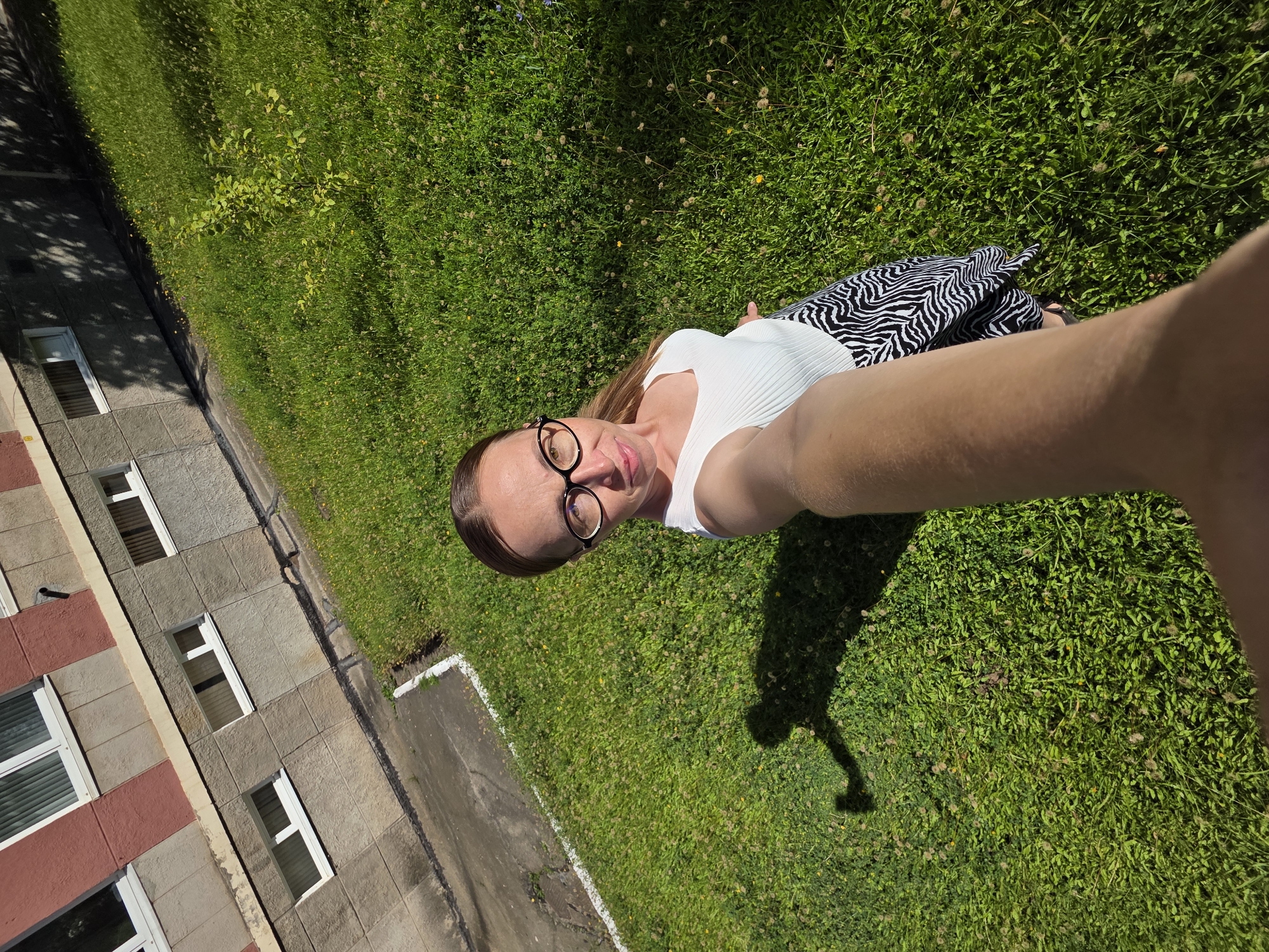 In Chaikovsky, Russia, a woman with light brown hair pulled back and wearing glasses, a white sleeveless top, and a black and white zebra-print skirt is taking a selfie outdoors. She is standing on a grassy area with patches of clover and small yellow wildflowers. Behind her is a concrete sidewalk with a white border and a multi-story building with grey and red sections and white-framed windows. The woman's shadow is visible on the grass, cast by the sunlight.