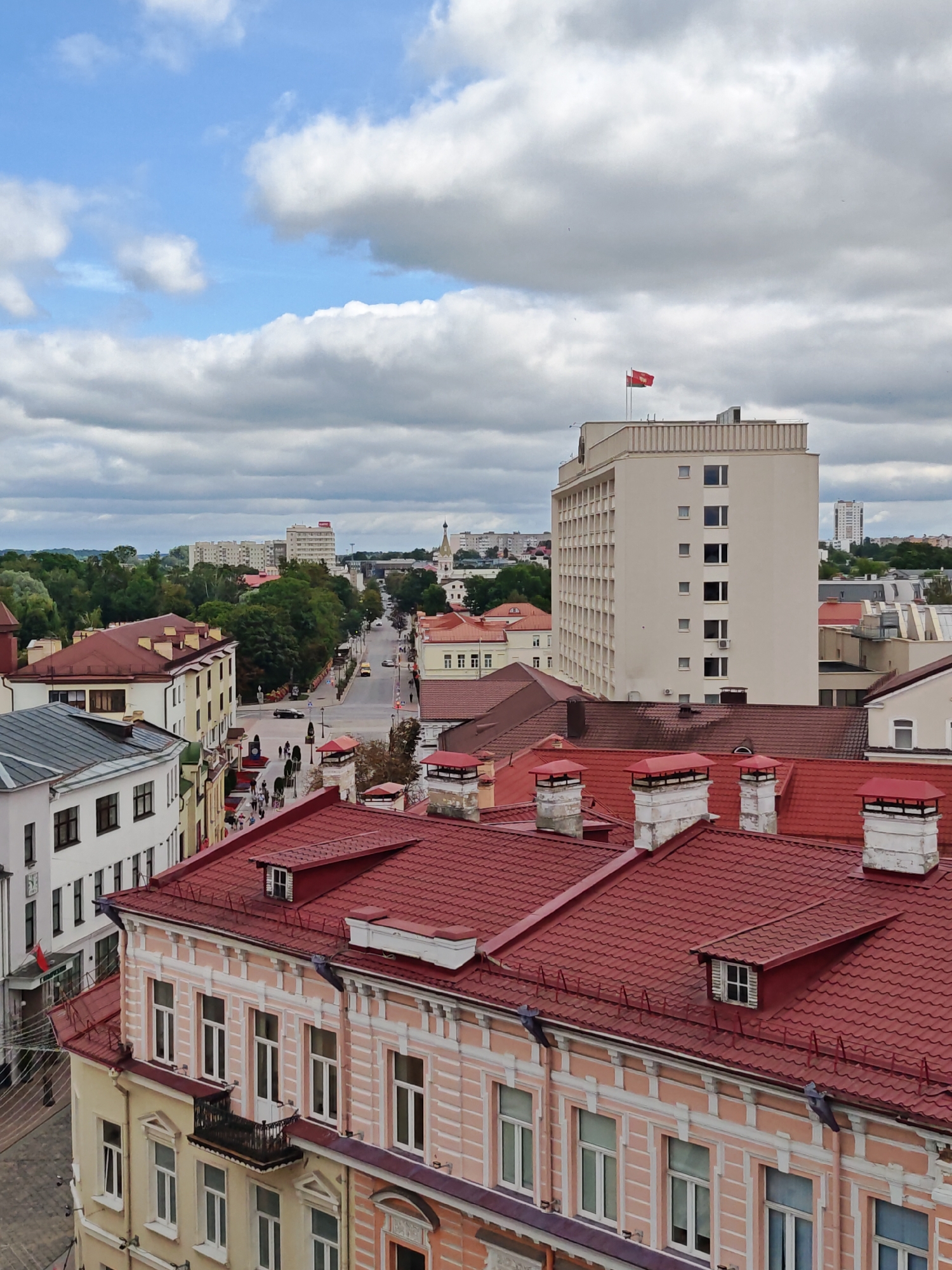 This is an aerial, full-frame, vertical shot of a city street in Hrodna, Belarus, with buildings and a cloudy sky. In the foreground, several red-tiled roofs of old buildings are visible. The buildings have light-colored facades with ornate white trim and windows. Further back, a wide street lined with trees and more buildings stretches into the distance. Pedestrians are visible walking along the sidewalk. A tall, modern, beige building with many windows stands prominently in the mid-ground, with a red and yellow flag flying from its rooftop. The sky is filled with a mix of blue sky and large, white, fluffy clouds.
