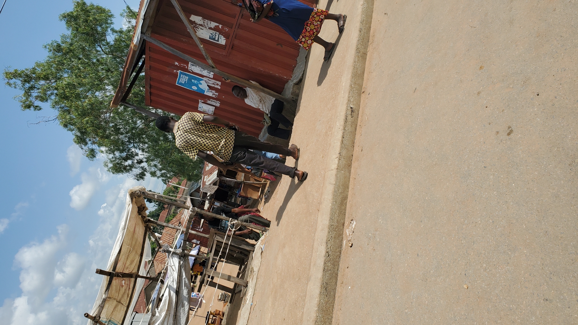 A medium shot captured outdoors on a sunny day depicts a street scene in Jalingo, Nigeria, characterized by informal market stalls and structures. In the foreground, a person wearing a black and yellow checkered shirt and dark pants stands facing away from the camera, casting a distinct shadow on the dusty ground. To their right, a man in a light-colored shirt and dark pants is seated, leaning against a corrugated metal shipping container painted red. To the far right, a person wearing a blue dress with a patterned hem is visible, also facing away from the camera. Behind these individuals, various market stalls are set up with white tarpaulin roofs supported by wooden poles, displaying goods and merchandise. The background features a large, leafy green tree and a blue sky with scattered white clouds. The overall environment suggests a bustling, open-air market area within the city.