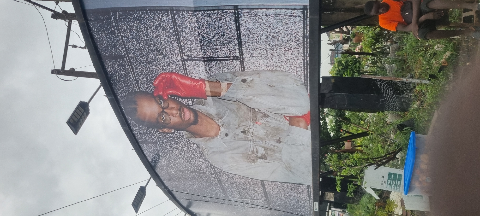 A billboard featuring a man wearing glasses and a red glove is displayed in the city of Moba, Nigeria. The man is dressed in a denim jacket and is touching his face with the gloved hand. The billboard is mounted on a metal frame, with street lights visible above it. In the foreground, there is a young man sitting on a chair, wearing an orange t-shirt and black shorts. He is holding a bag on his lap. Behind him, there are various plants and a blue container filled with oranges. A fence made of metal wire encloses the area. The sky is overcast.