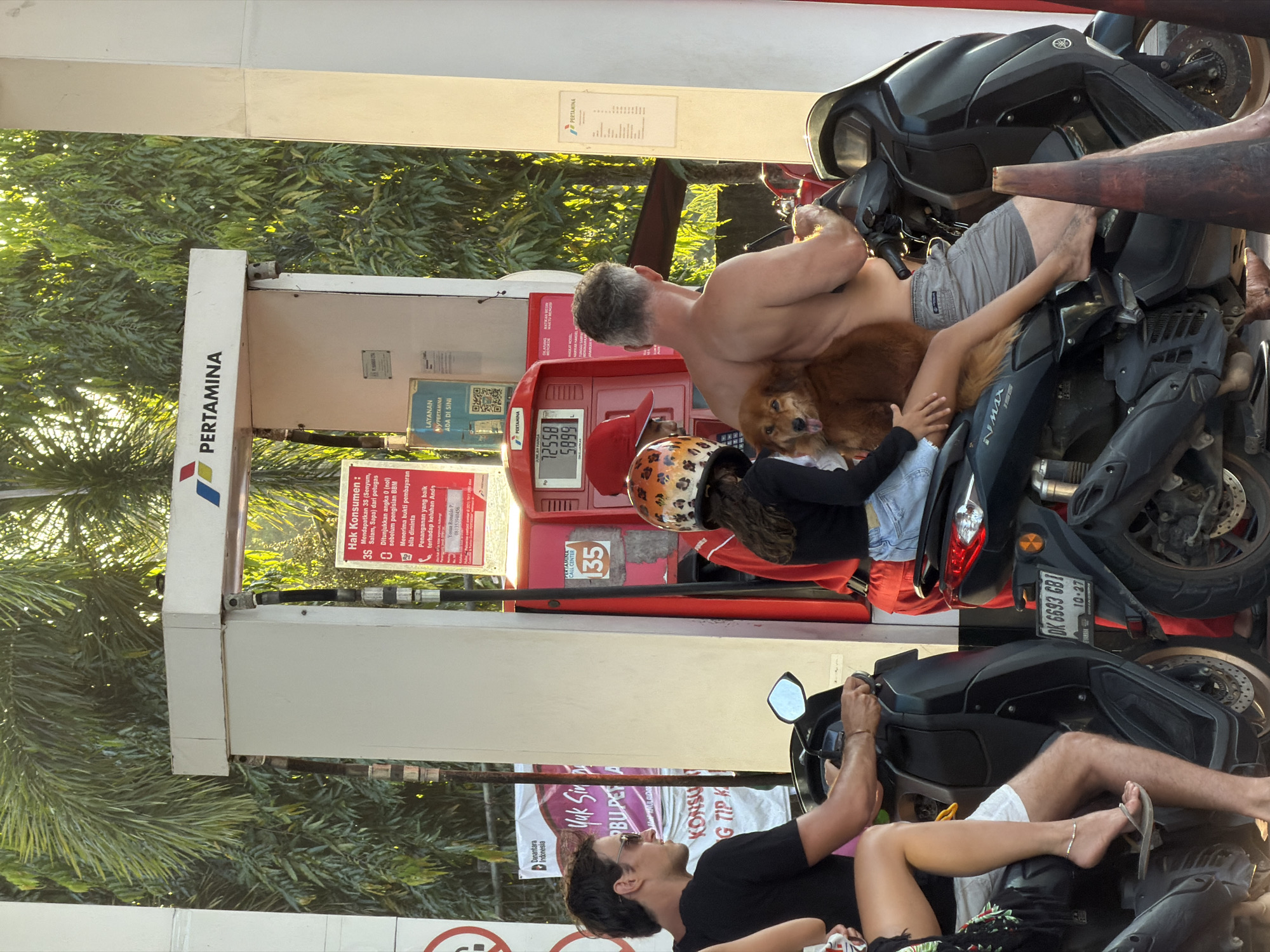 The image depicts a gas station in Mengwi, Indonesia. The station is branded with "PERTAMINA". A shirtless man is sitting on a black scooter, holding a brown dog. A child wearing a leopard-print helmet is also on the scooter. Another man is on a separate, identical black scooter, with a woman sitting behind him. The scooters are positioned at the gas pumps. The gas pump's display shows a price of 5899 and a volume of 72558. Signs on the gas pump are in Indonesian and include the logo of Pertamina. A license plate visible on one of the scooters reads "DK 6693 GBI" and expires on "10-27". Foliage is visible in the background.