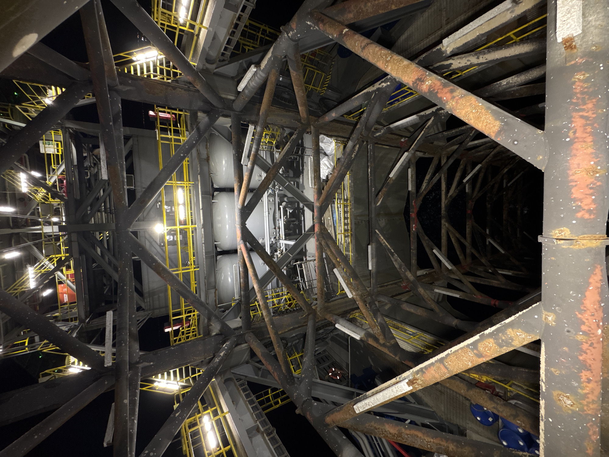 The image shows a perspective looking up into the complex metal framework of what appears to be an industrial structure, possibly a drilling rig or a large tower, at night. The structure is made of dark, weathered metal beams and supports, many of which show signs of rust and wear. Yellow railings and staircases are visible at various levels within the framework, illuminated by numerous bright white lights. These lights create strong highlights on the metal surfaces and cast shadows. In the background, the dark night sky is visible, with the lights of a distant city faintly appearing on the horizon. Several large, cylindrical white tanks are also part of the structure. Two blue barrels are partially visible in the lower left portion of the image. The overall impression is of a functional, utilitarian structure designed for industrial purposes, viewed from a low angle looking upwards towards its upper reaches. There are no people or animals visible in the image. The location is described as "Unknown, Italy" in the prompt.