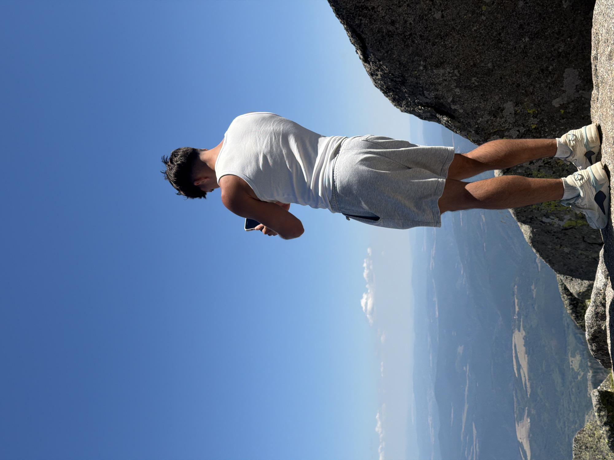 A person is standing on a rocky outcrop on a mountain, taking a picture with their phone. The person is wearing a white tank top and gray shorts. The background is a clear blue sky and a vast, mountainous landscape. The location is on Mount Vitosha near Bistritsa, Bulgaria.