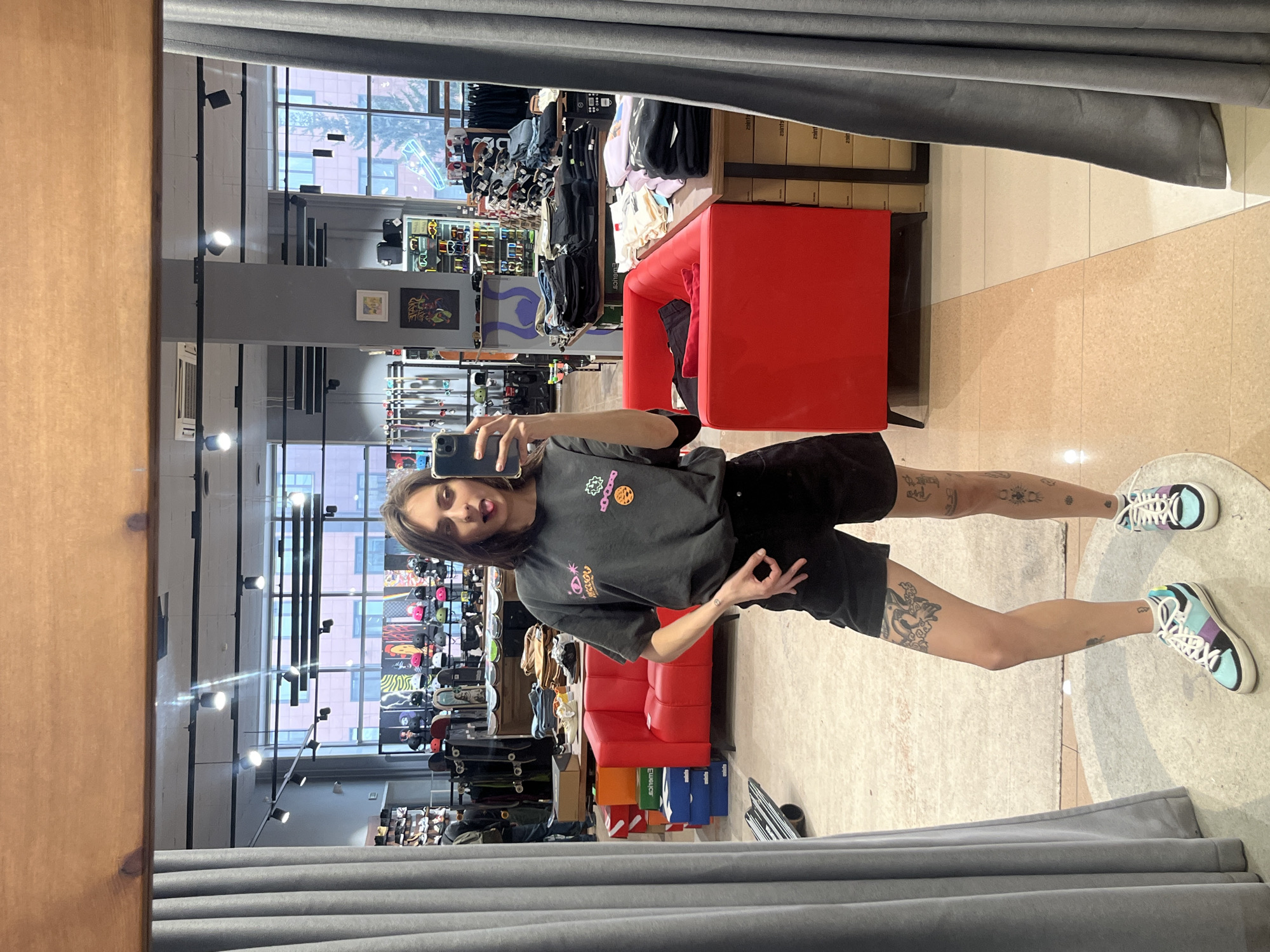 A person with light skin, a short hairstyle, wearing a dark t-shirt with colorful cartoon designs, and dark shorts is reflected in a mirror inside a retail store. The individual is holding a phone, presumably taking a picture, and is making an "OK" hand gesture. The store has rows of merchandise, including skateboards and shoes. Behind the individual in the reflection, there is a red seating area. The location is identifiable as the "Sneaker Head" store at Mira 91 in Krasnoyarsk, Russia.