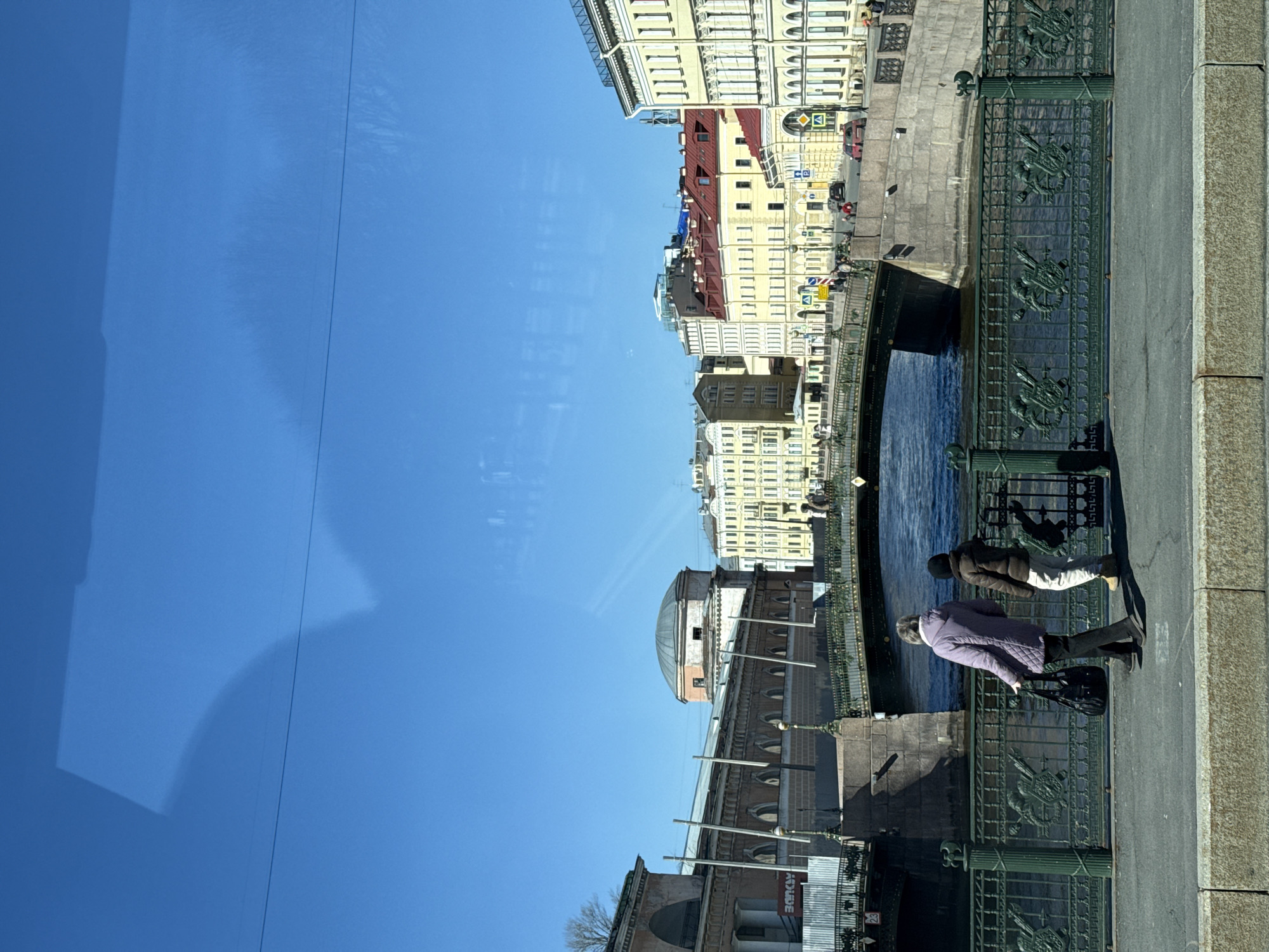 The image shows a street scene in Saint Petersburg, Russia, likely captured from a moving vehicle. The viewpoint is near the Green Bridge (Zeleny Most), with a partial reflection of the vehicle visible in the frame. Two individuals are present on the bridge sidewalk; one wears a purple coat and carries a black bag, while the other wears a brown jacket and light-colored pants. They appear to be observing something below the bridge. The fence of the Green Bridge is visible, decorated with ornate metalwork. In the background are multi-story buildings and other urban infrastructure. The sky is blue and clear. The angle of the photo is vertical, so the image is rotated ninety degrees.