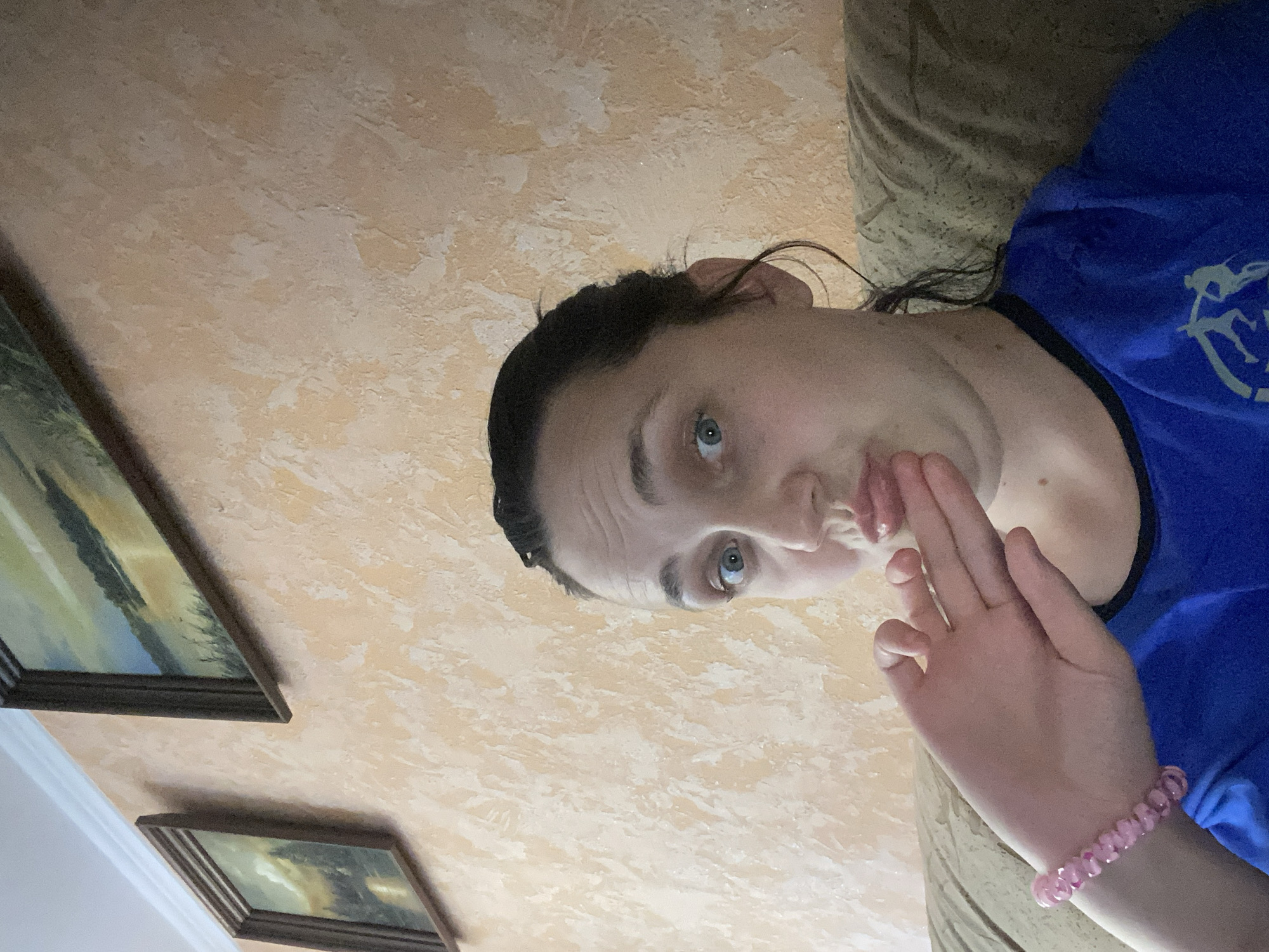 An individual is taking a selfie indoors, sitting on a couch against a textured, peach-colored wall. The individual, with dark hair pulled back and eyes wide open, is looking directly at the camera with their lips pursed. Their left hand is raised to their mouth, with a pink beaded bracelet on their wrist. The individual is wearing a blue t-shirt. Two framed landscape paintings hang on the wall behind them. The scene appears to be in a residential setting, possibly a living room.