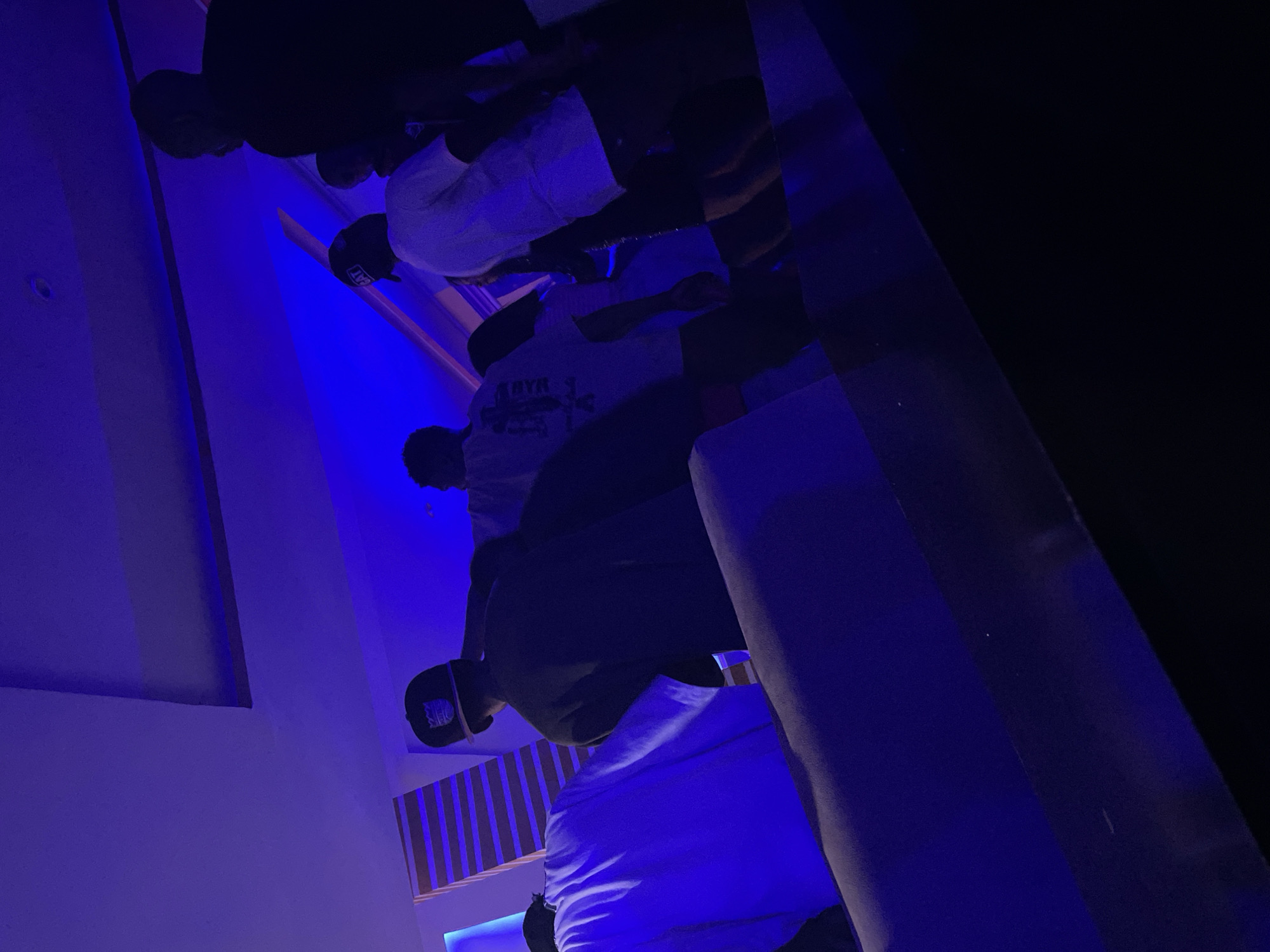 The image captures an indoor scene, primarily illuminated by intense blue LED lighting that bathes the left wall and casts a vibrant hue across the environment. The setting appears to be a dimly lit lounge, club, or entertainment space.
Several men are present, standing or sitting in a relaxed, social gathering. In the foreground, a man is visible wearing a black baseball cap with the word "CAT" clearly printed in white letters on the front, paired with a white t-shirt. Behind him, another man wears a white t-shirt that displays a dark graphic and visible text, possibly "ARYA." Further back, additional male figures are partially visible, mostly silhouetted against the dark background, also appearing to wear caps and light-colored tops.
The left side of the room features a light-colored wall, intensely lit by the blue LEDs. On the right, dark, possibly reflective surfaces and structured elements, potentially seating or part of the room's interior design, are discernible. A wall in the mid-ground displays a pattern of vertical stripes, creating visual interest. The ambient darkness combined with the artificial lighting strongly suggests the activity is taking place at nighttime. The overall atmosphere conveys a casual social event. This scene is set in Durumi, Nigeria.